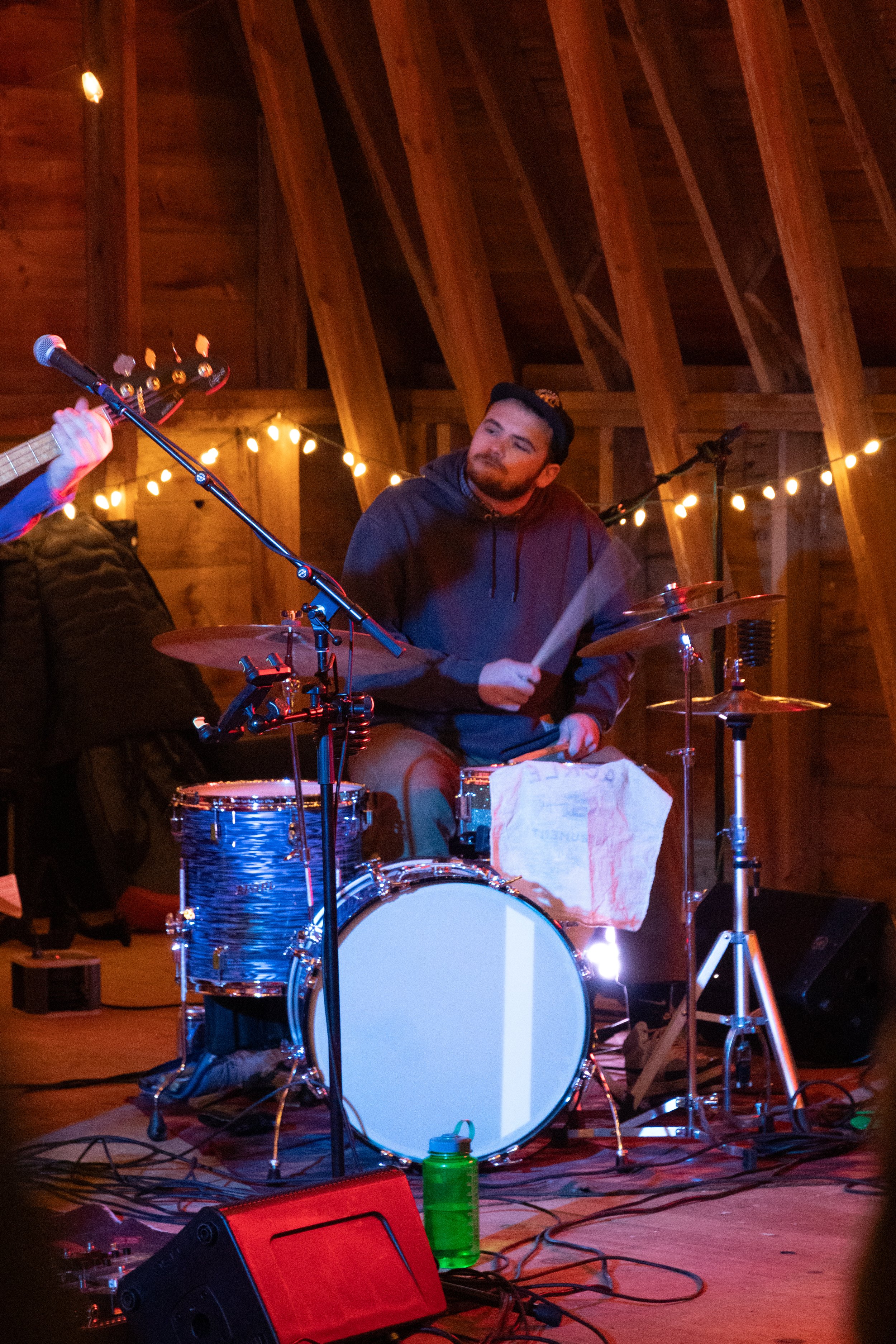A man playing a drum set during a live performance in a wooden venue with string lights.