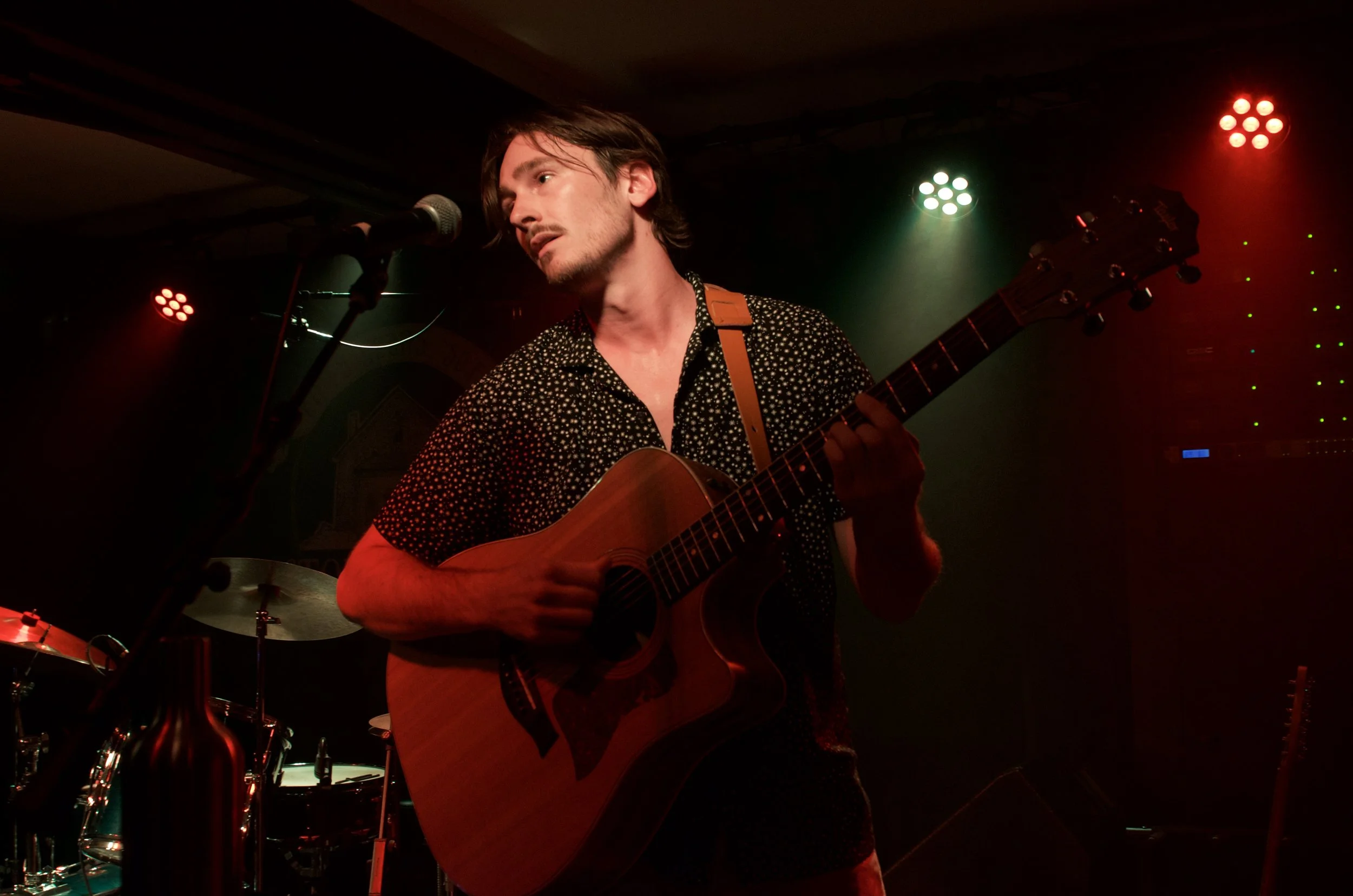 A young man playing an acoustic guitar on stage, singing into a microphone, with colorful stage lights overhead.