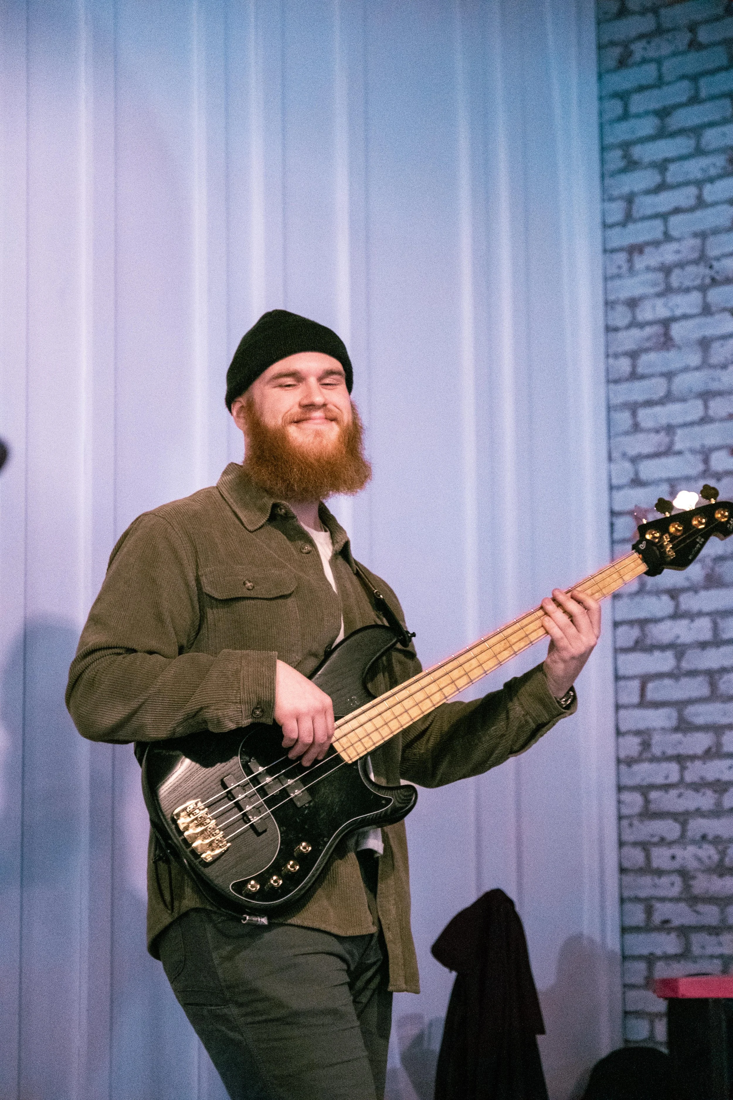 A man with a beard wearing a black beanie and brown jacket playing a black bass guitar, smiling with eyes closed in an indoor setting with white and gray brick walls.