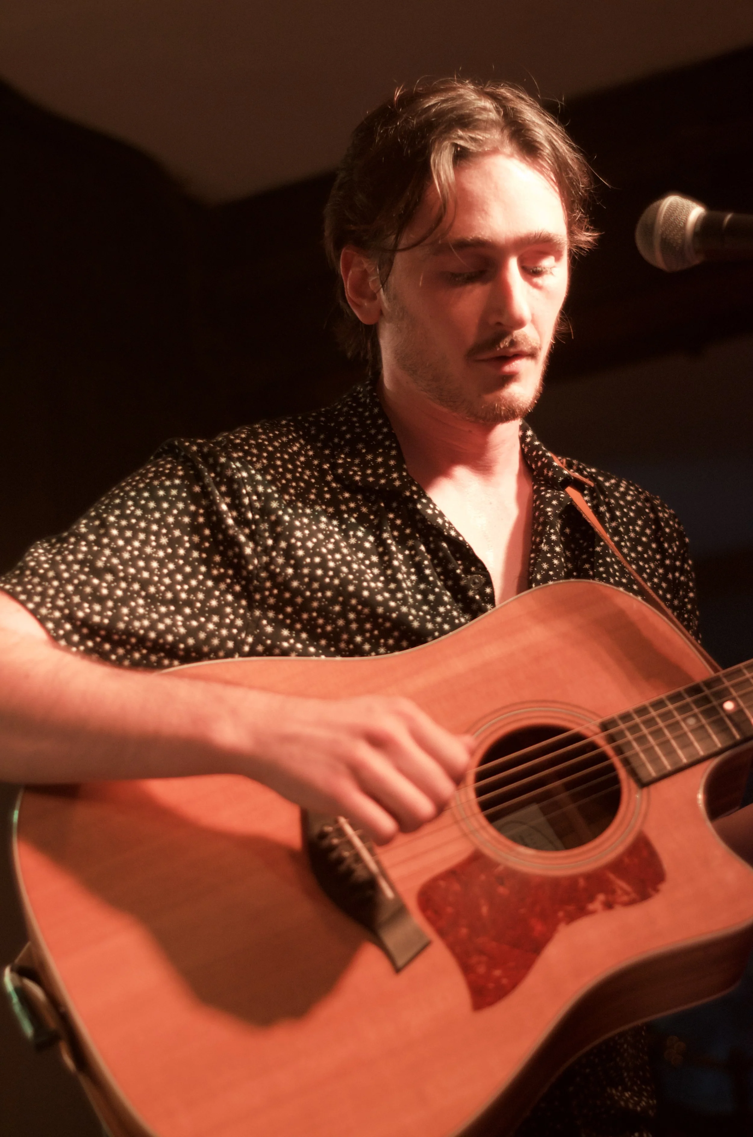 A young man playing an acoustic guitar on stage, wearing a black shirt with white stars, standing in front of a microphone with warm stage lighting.