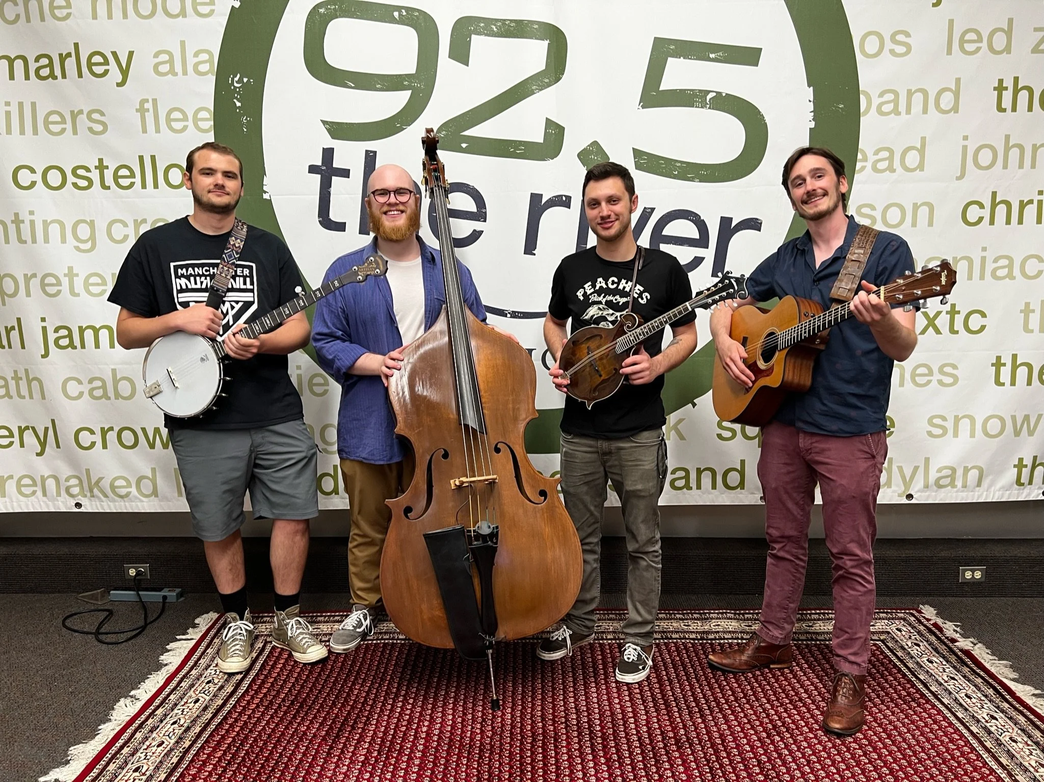 Four men standing with musical instruments in front of a large sign reading '92.5 the river'. The first man on the left holds a banjo, the second man holds a double bass, the third holds a mandolin, and the fourth holds an acoustic guitar. They are smiling and standing on a red patterned rug.