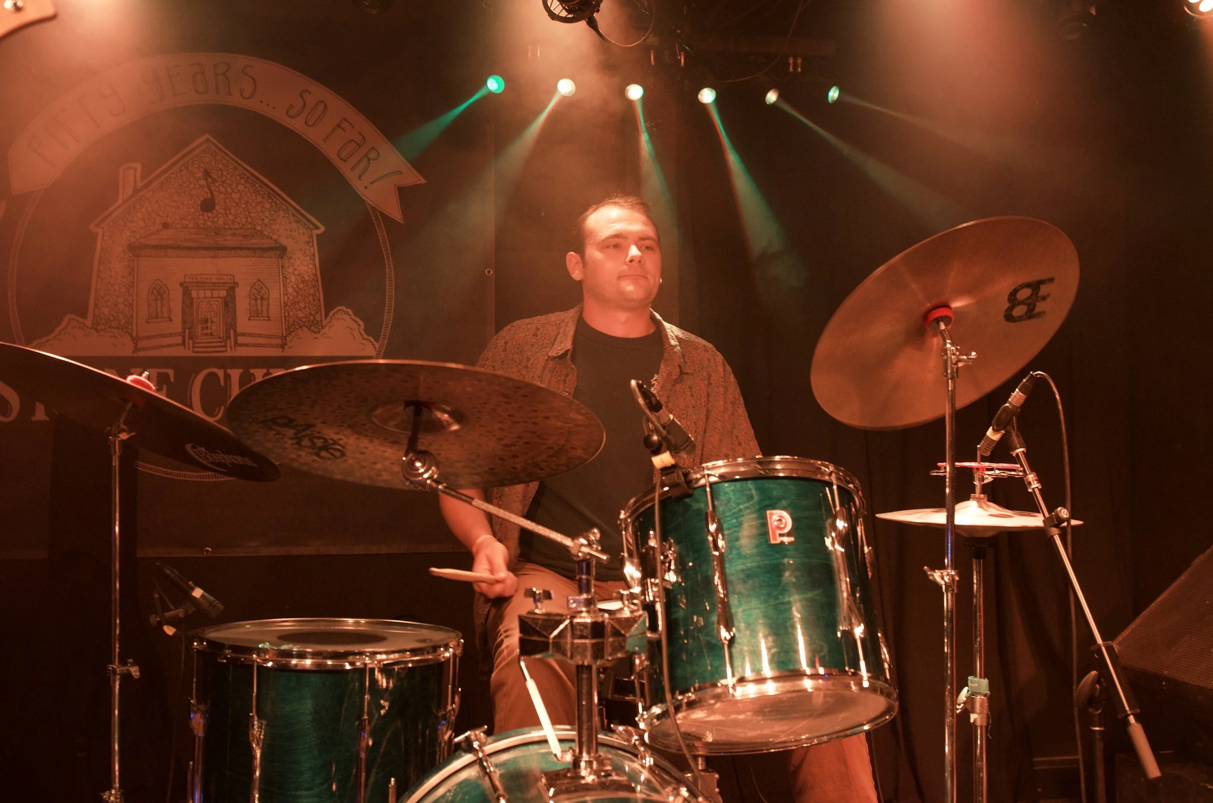 A male drummer playing drums on stage during a live performance, with stage lights and a backdrop.