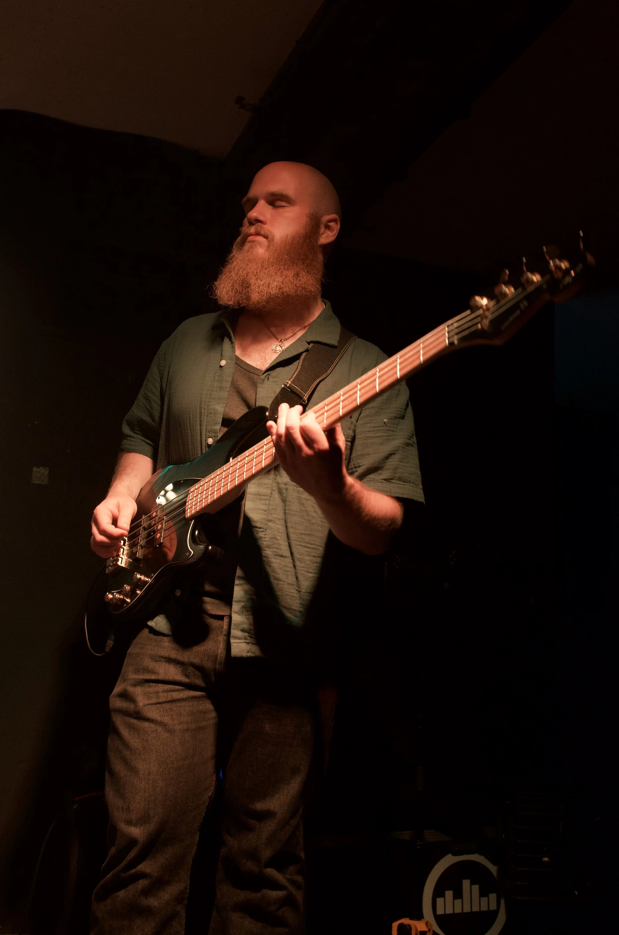 A bearded man with a shaved head playing an electric bass guitar on stage with a dark background.