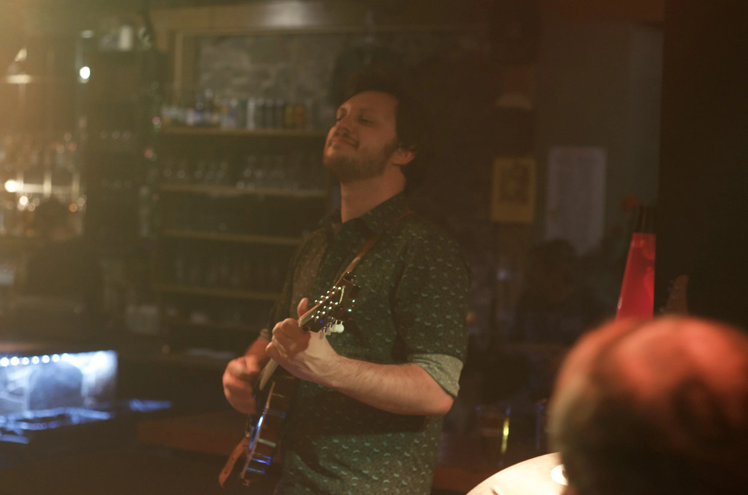 A man playing an electric guitar in a dimly lit venue, with shelves of bottles and glasses in the background.