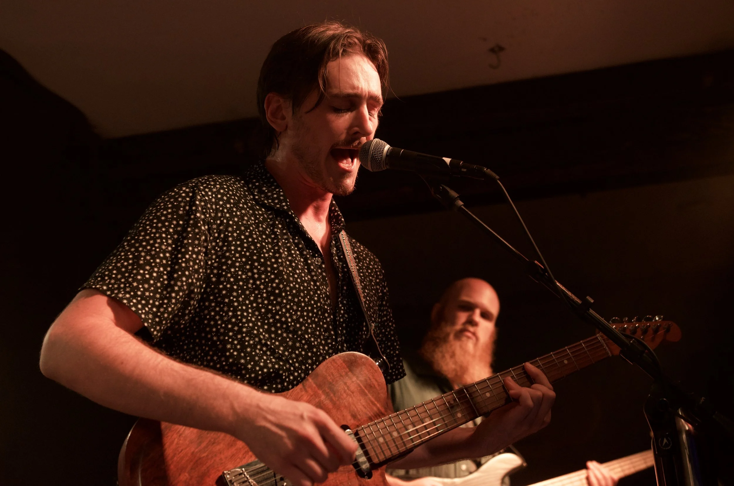 A man with brown hair singing into a microphone while playing an acoustic guitar, with another man with a beard playing bass guitar in the background.