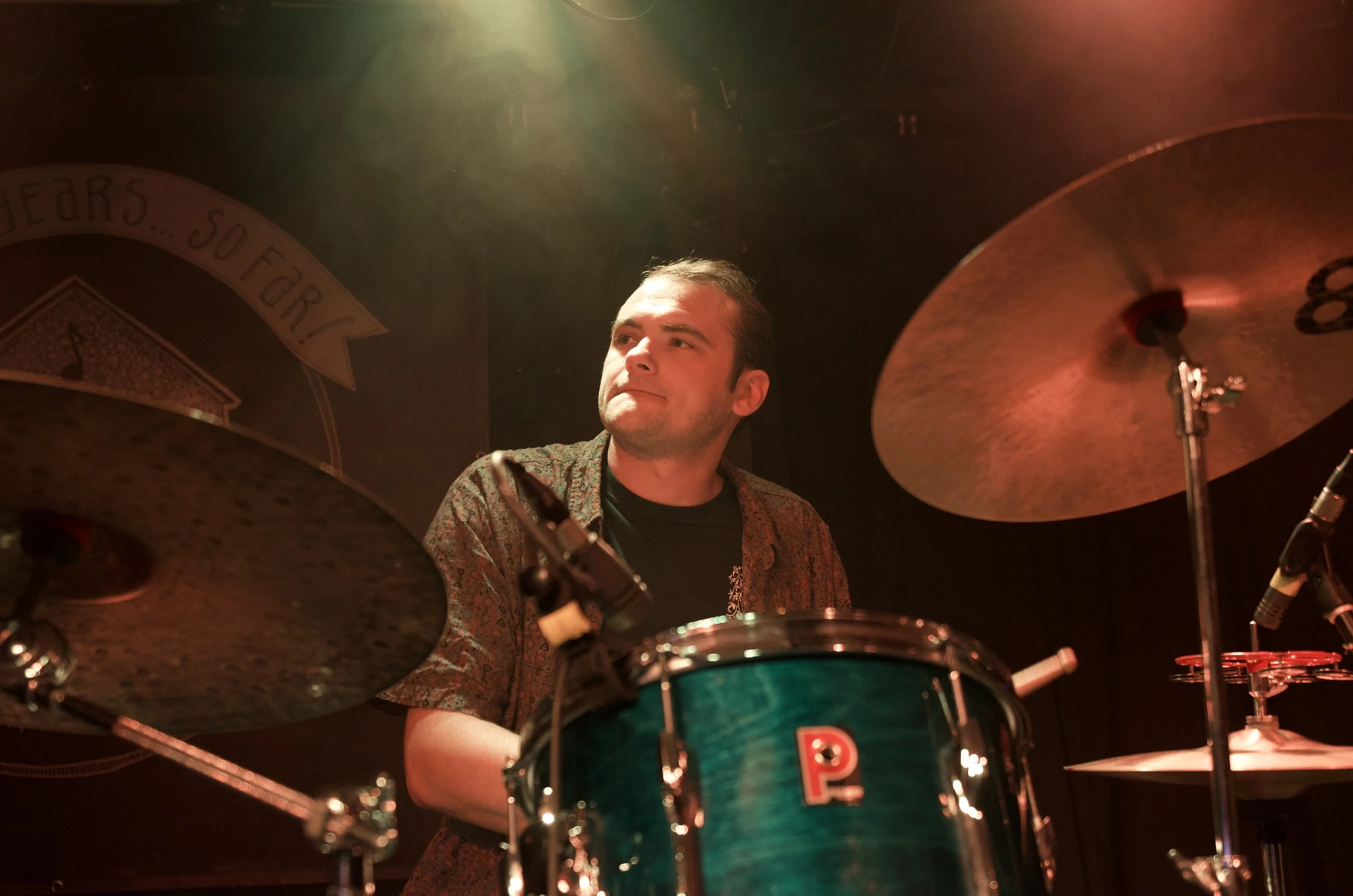 A man playing drums on stage, with cymbals on either side and a black backdrop.