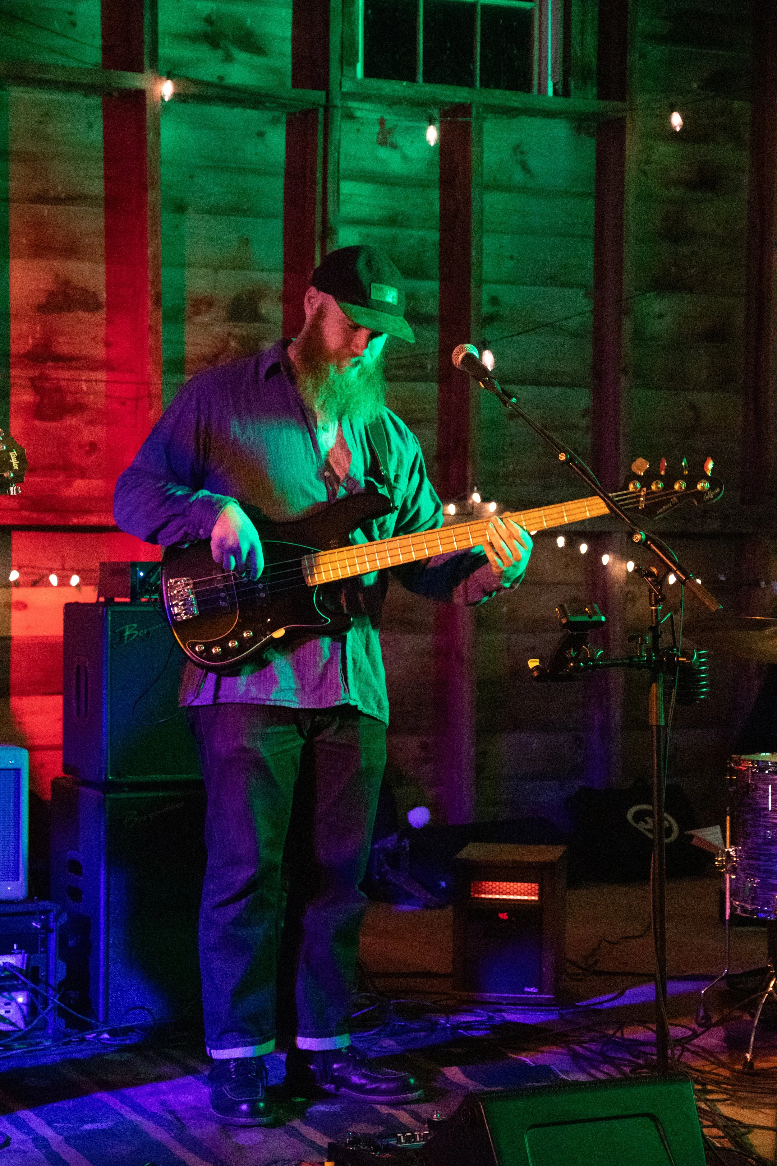 A musician with a beard and glasses playing an electric bass guitar on a stage with colorful lighting and a wooden background.