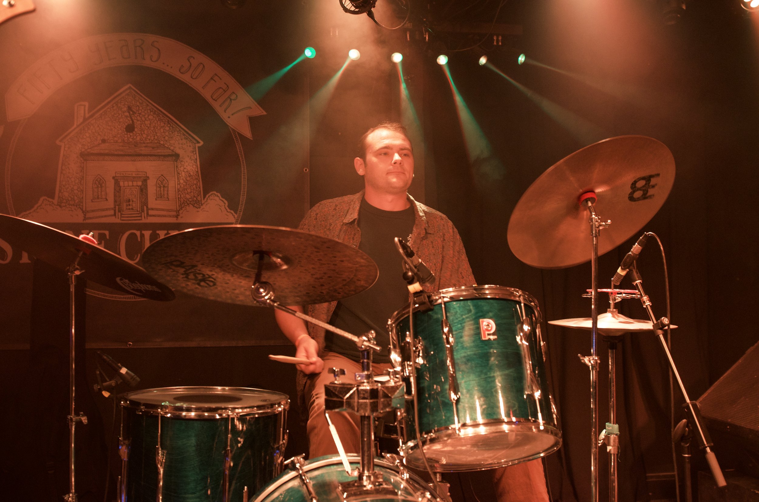 A man playing a drum set on stage with colorful lights and a backdrop featuring a house drawing and the text 'Fifty Years, So Far'.