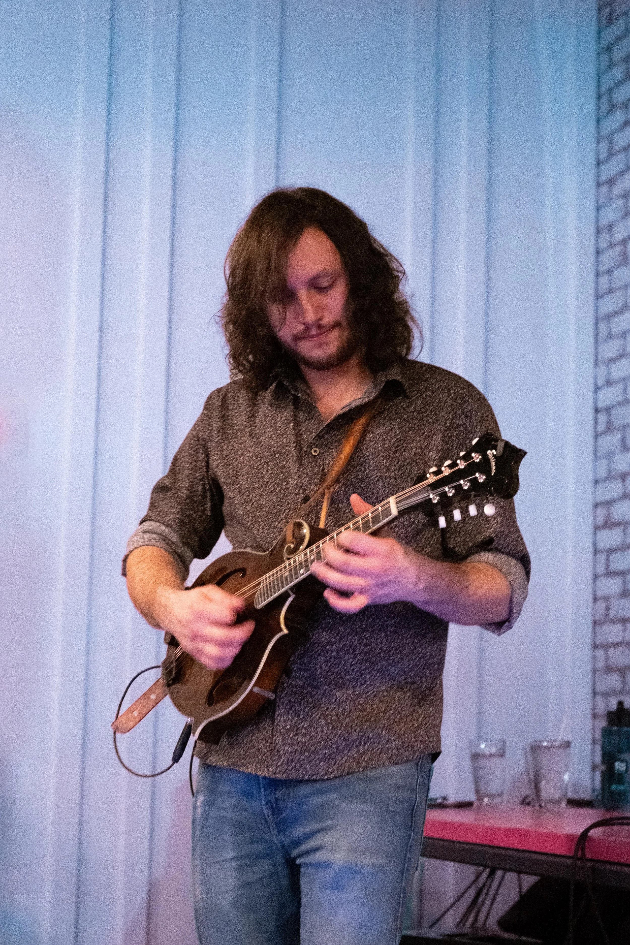 A man with long, curly hair playing a small stringed instrument, possibly a mandolin or ukulele, in a room with light blue panel walls and a part of a brick wall visible on the right.