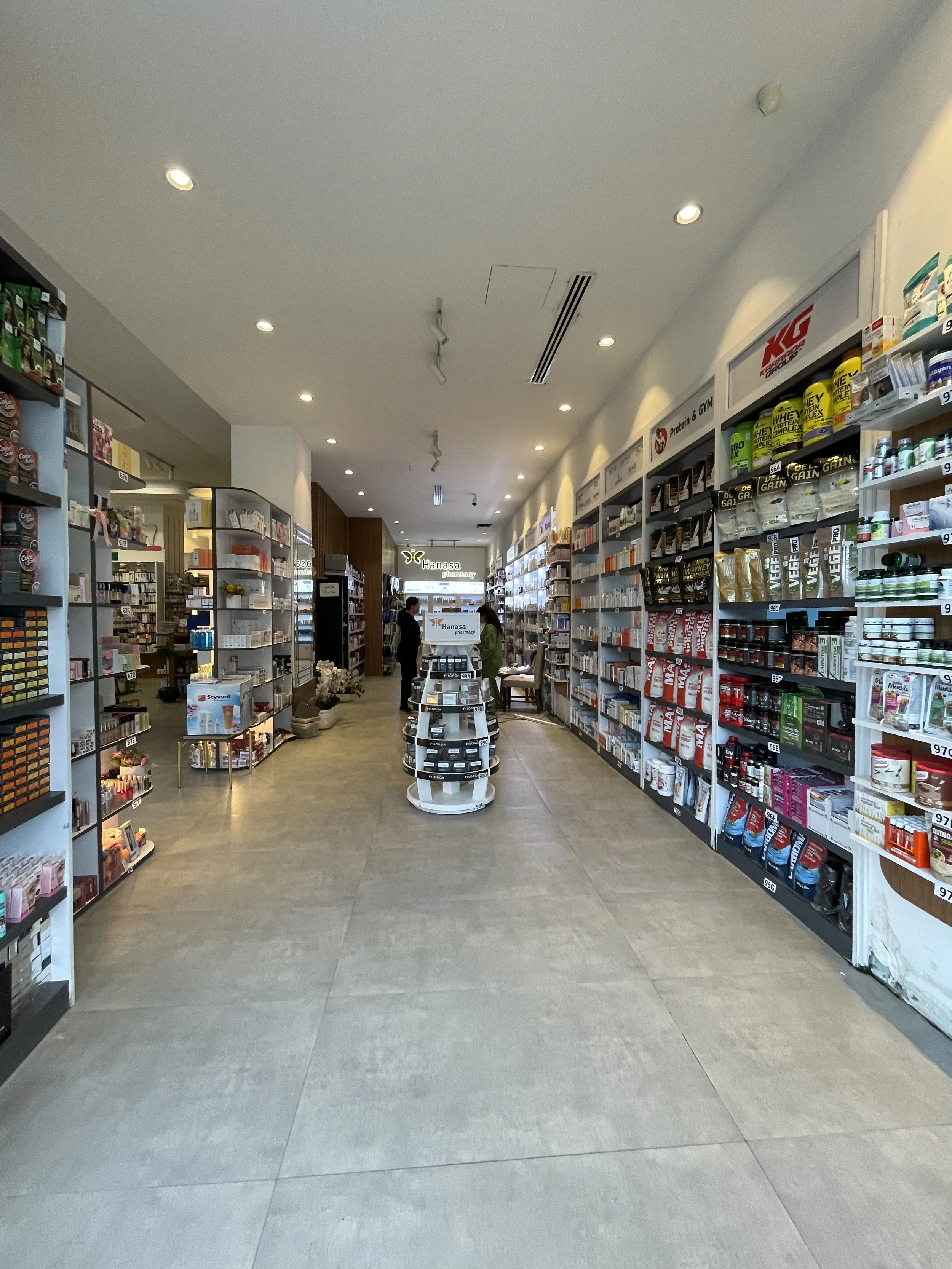 Interior of a pharmacy or health store aisle with shelves stocked with vitamins, supplements, and health products, with a few customers browsing.