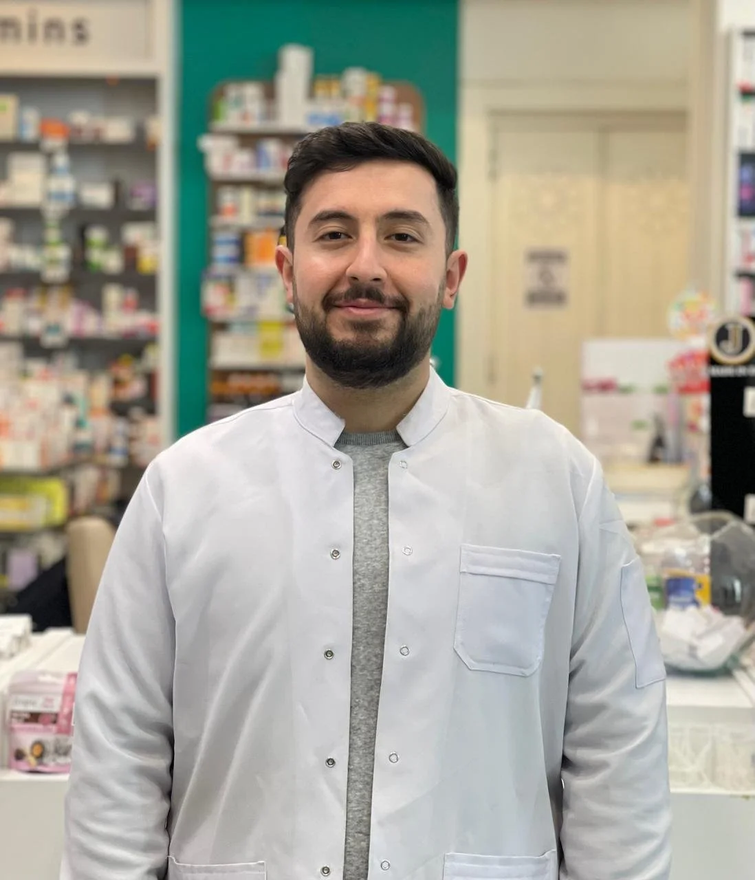A young man with dark hair and beard, wearing a white lab coat, standing in a pharmacy.