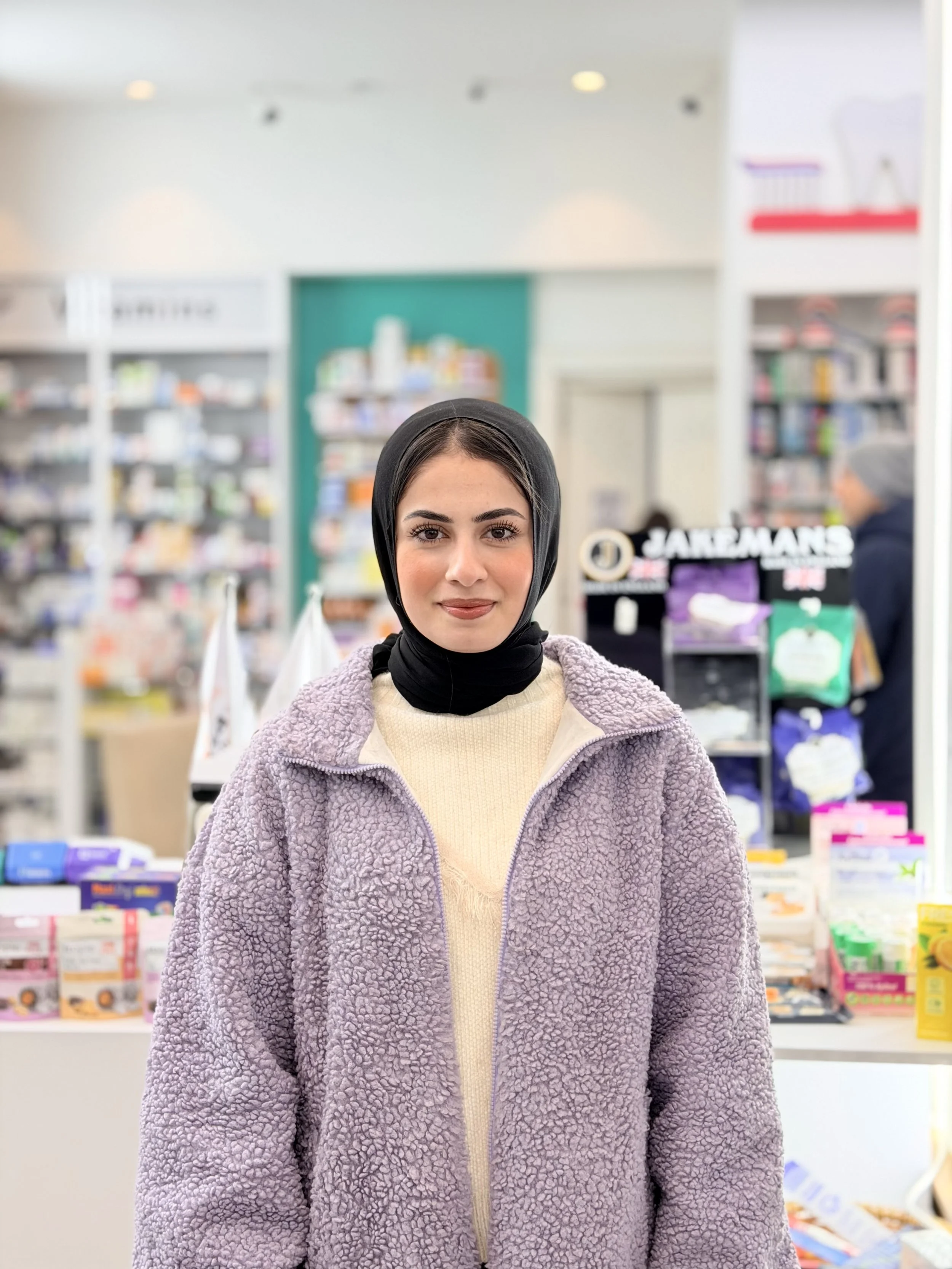 Young woman with dark eyebrows and black headscarf, wearing a cozy purple fleece jacket and cream sweater, standing inside a store with shelves of products in the background.