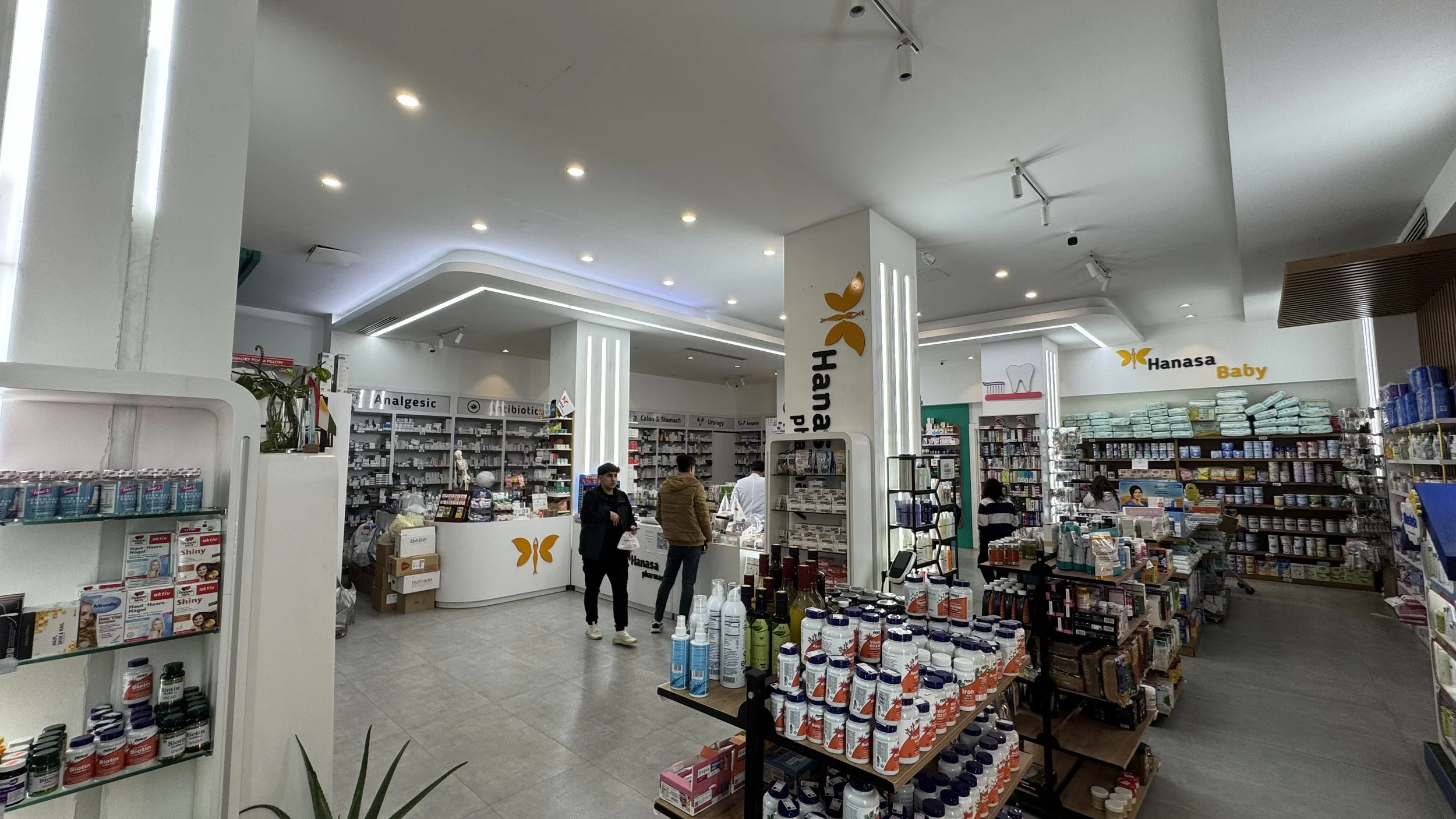 Interior of a pharmacy with shelves stocked with medicines and health products, and customers shopping.