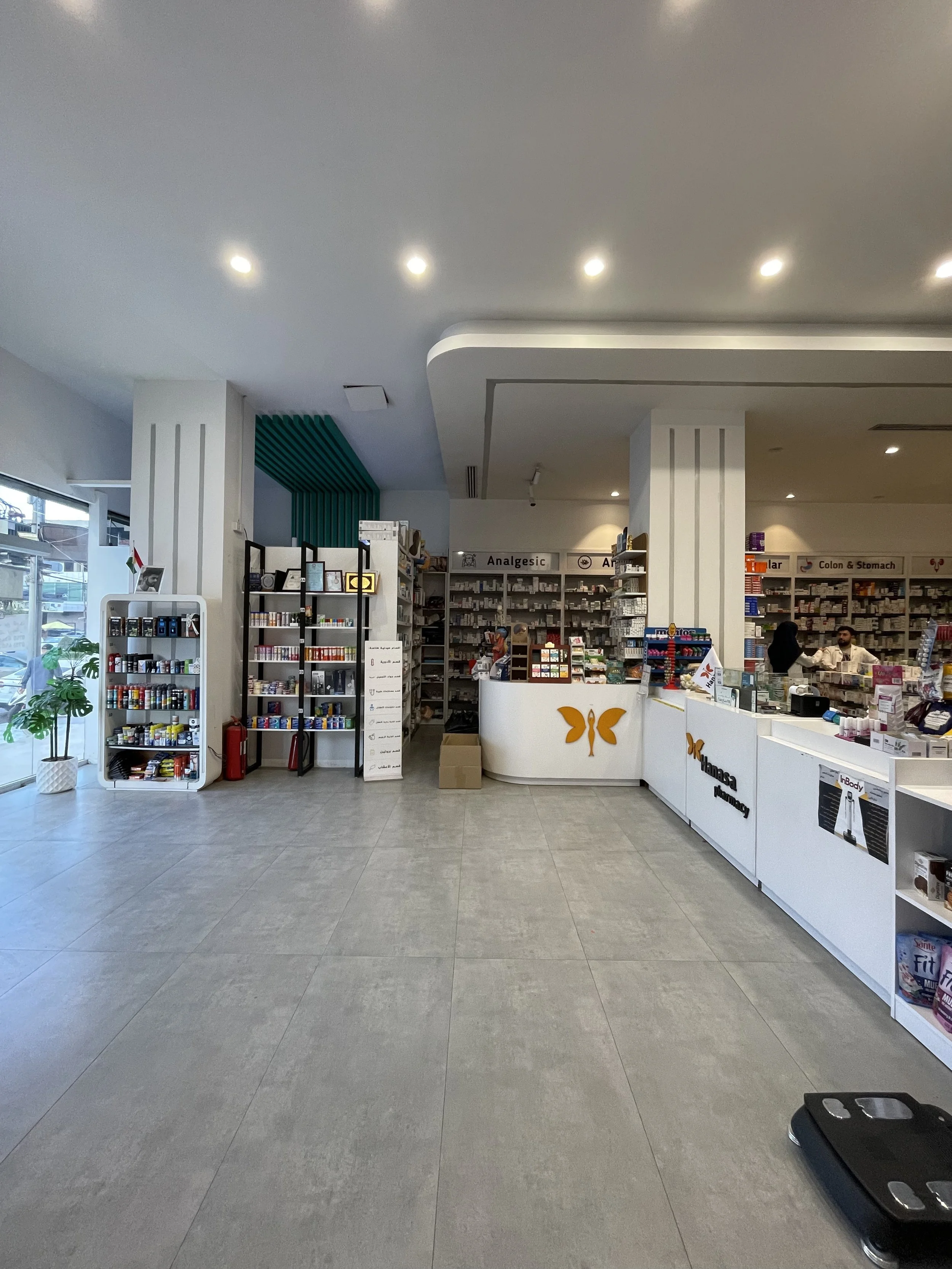 Interior of a pharmacy with shelves stocked with medications and health products, a curved white counter with pharmacy signage, and a scale on the floor.