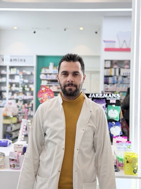 Man with dark hair and beard standing inside a pharmacy or drugstore, wearing a beige coat and mustard yellow turtleneck, with shelves of products in the background.