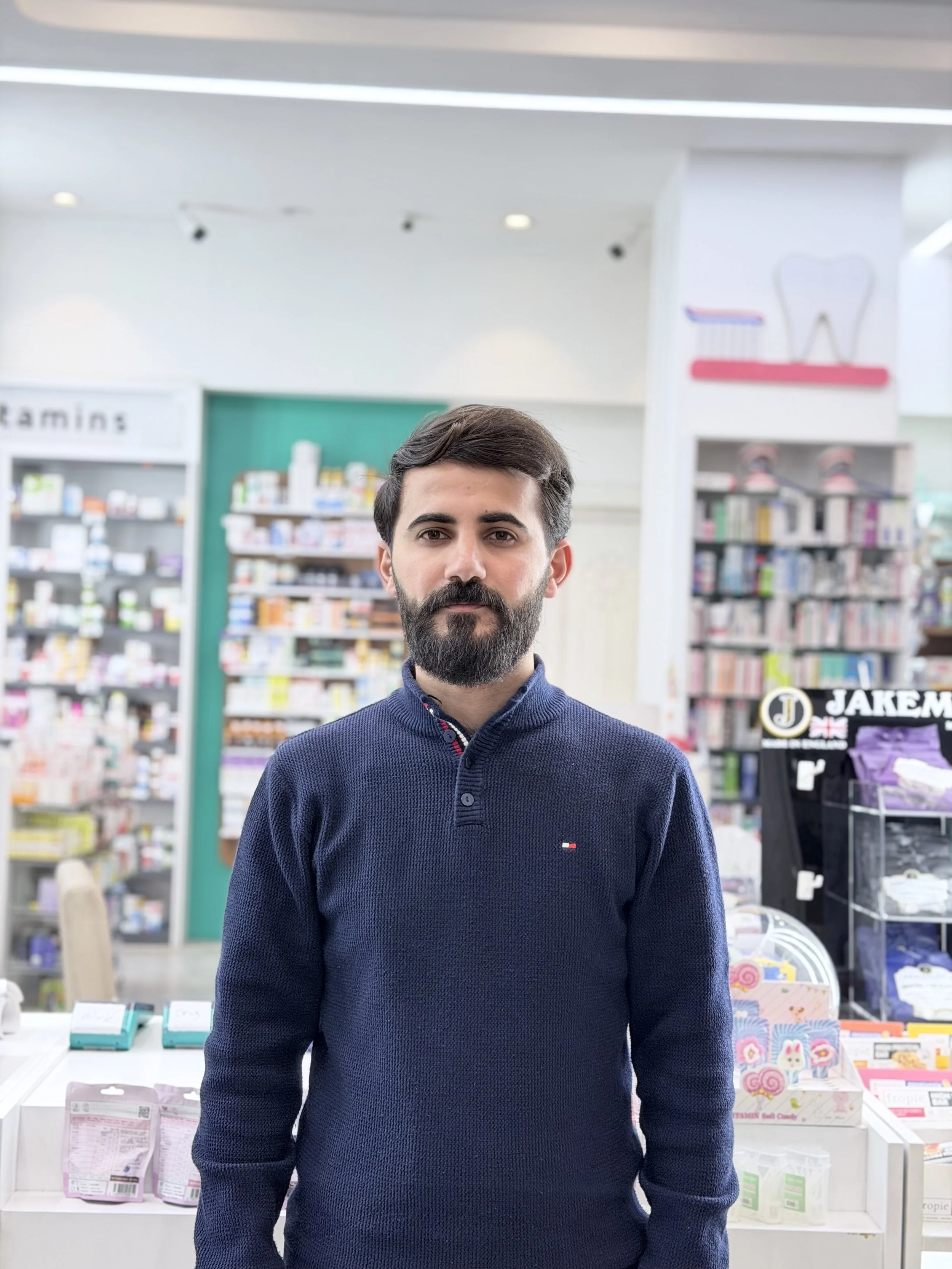 A man with a beard wearing a navy blue sweater standing in a pharmacy or health store with shelves of products in the background.
