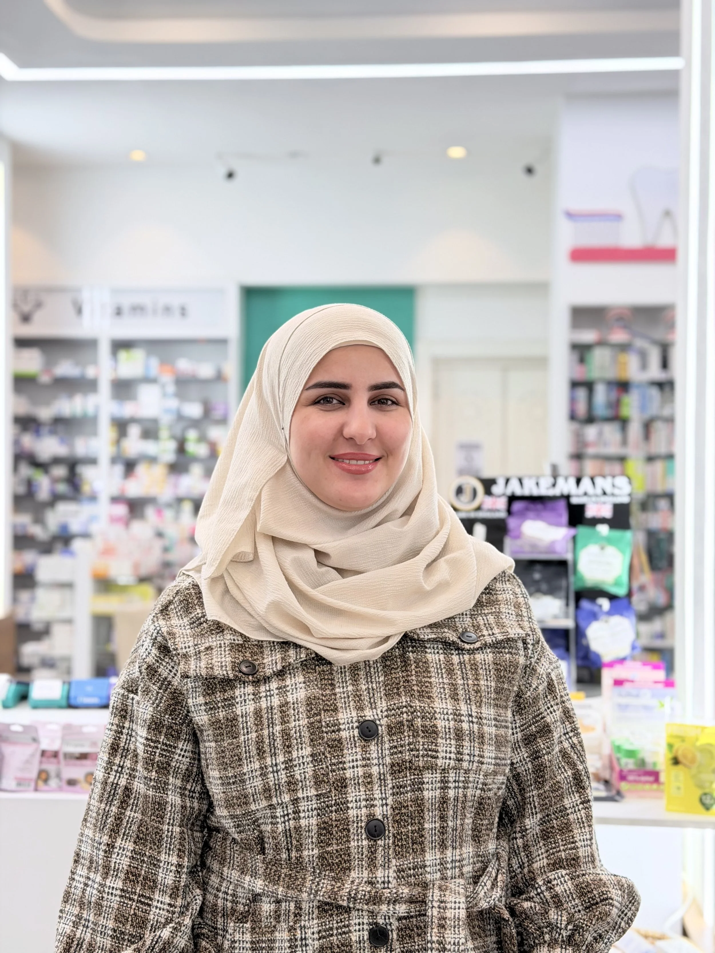 A woman wearing a beige hijab and a plaid coat, smiling in a store with shelves of products in the background.