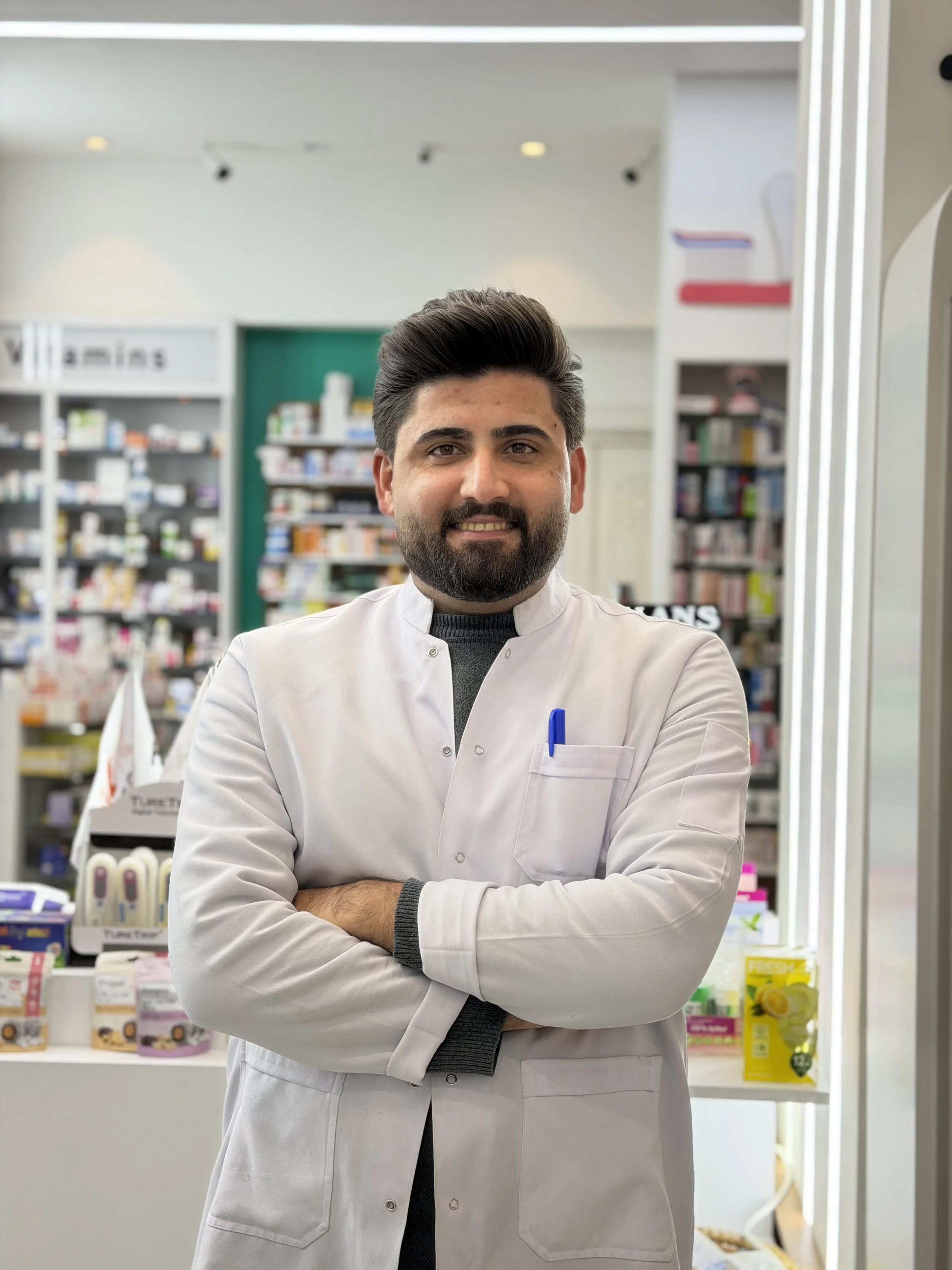 A male pharmacist with dark hair and beard standing cross-armed inside a pharmacy, with shelves of medicines and health products in the background.