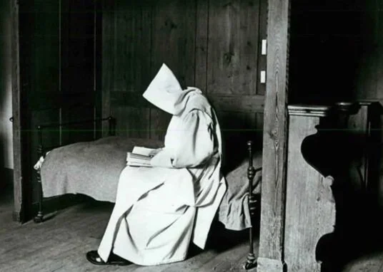 A person wearing a hooded robe sitting on a chair next to a bed, reading a book in a wood-paneled room.
