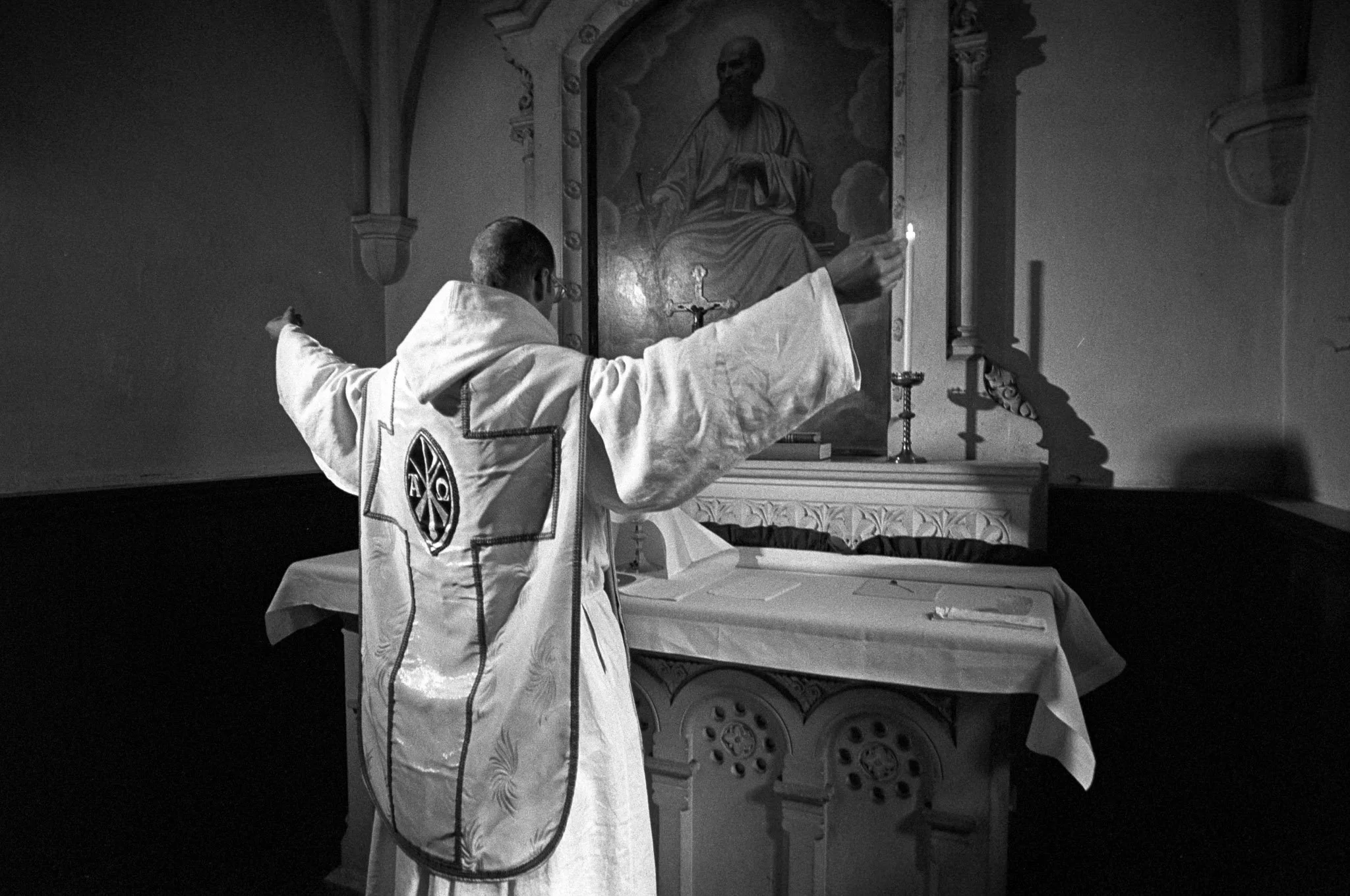 A priest in liturgical vestments lighting a candle in front of a religious painting, inside a church.
