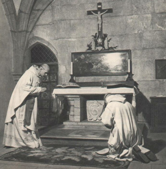 Two monks kneel and pray in front of an altar with a crucifix inside a stone church.