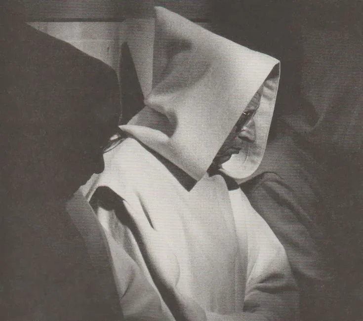 A person wearing a hooded outfit with a face covering, possibly depicting a nun or a person in costume, sitting next to another person with dark hair and glasses in a black-and-white photo.