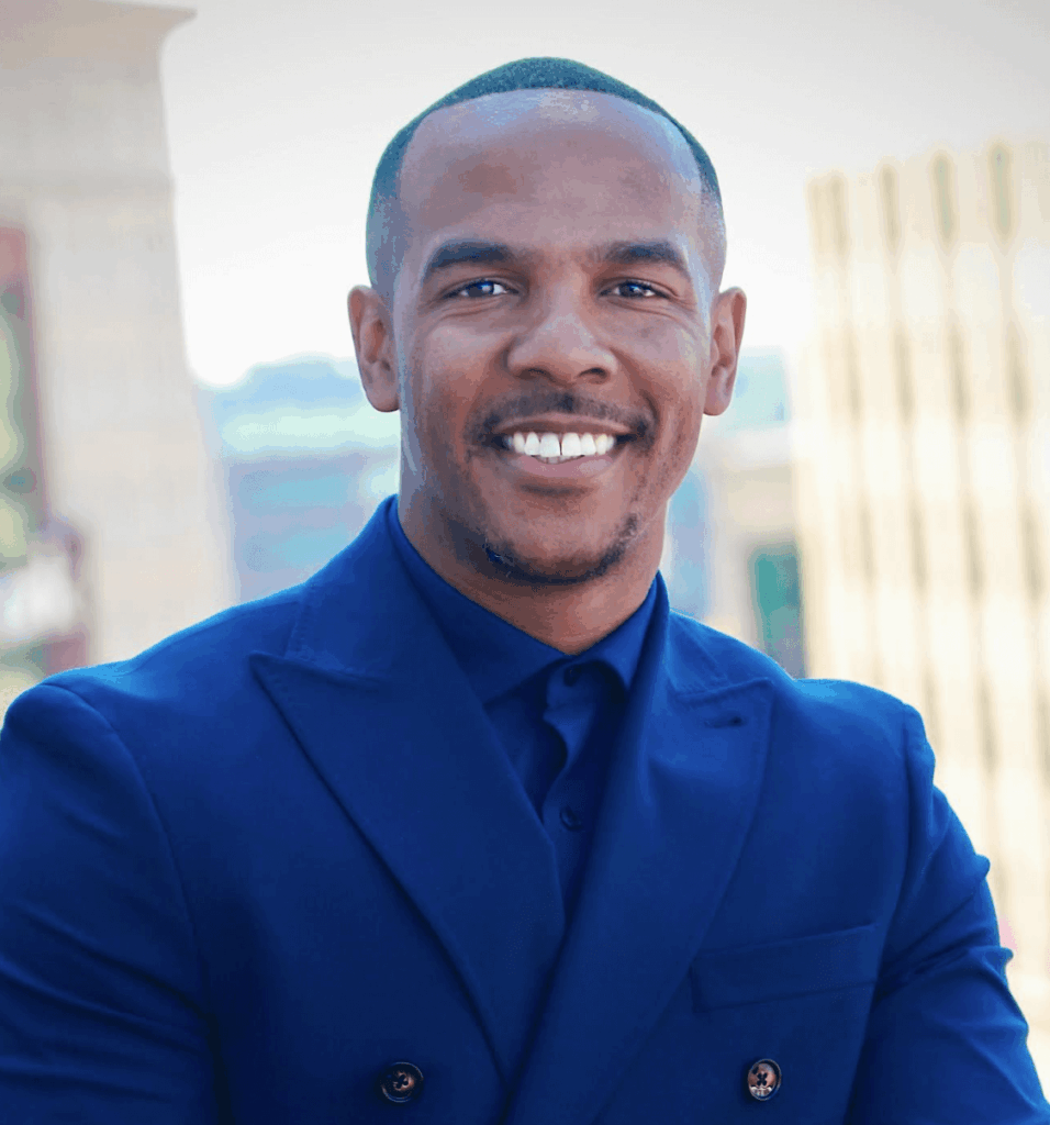 A smiling young man with dark skin, short black hair, wearing a bright blue suit and shirt, standing outdoors with a blurred cityscape background.