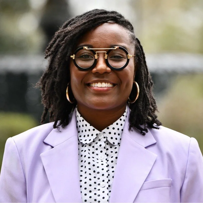 A smiling young man with dark skin, short black hair, wearing a bright blue suit and shirt, standing outdoors with a blurred cityscape background.