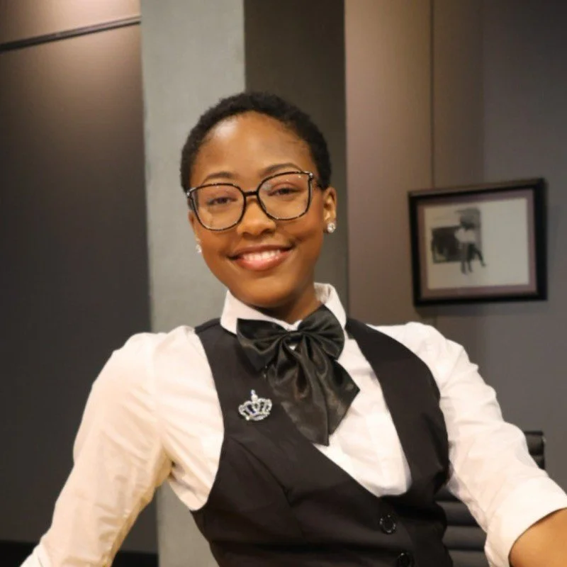 A woman in a formal uniform with glasses, a bow tie, and a brooch, smiling in an indoor setting with framed art on the dark-colored wall behind her.