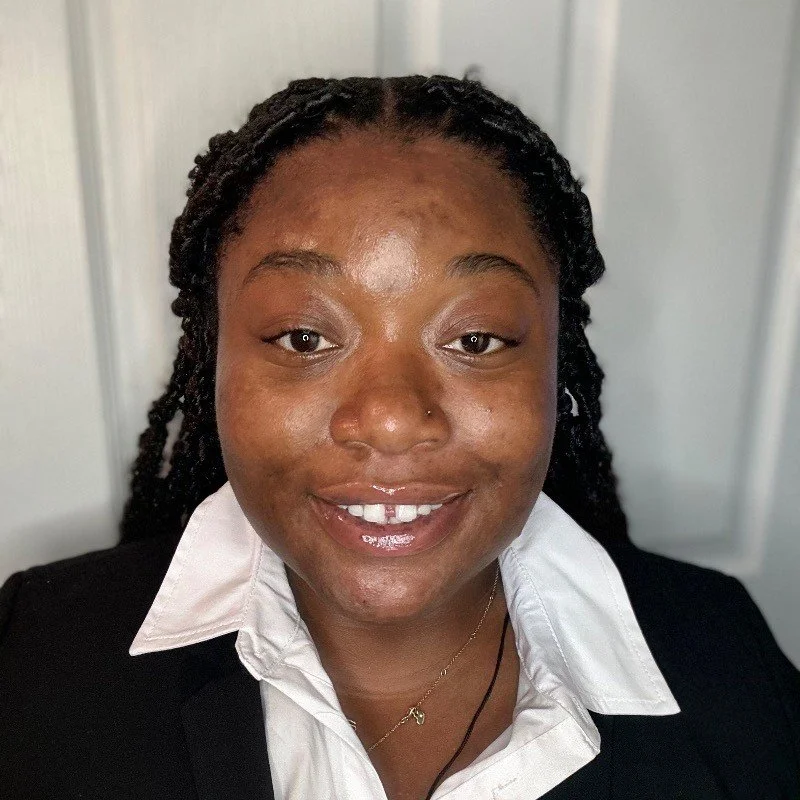A woman with braided hair, wearing a white collared shirt and black blazer, smiling in front of a light-colored background.