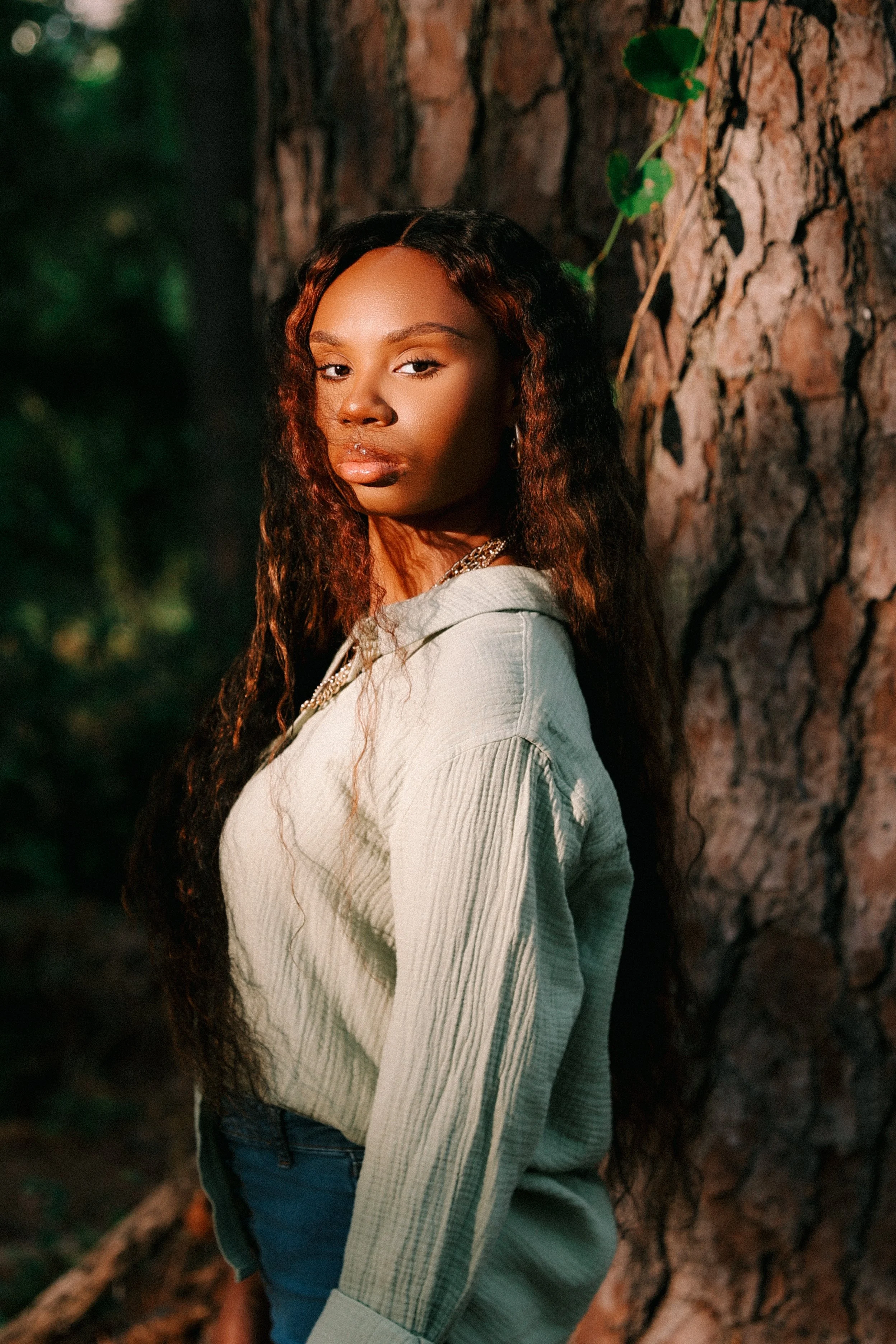A young woman with wavy hair posing outdoors against a tree trunk, looking at the camera. She is wearing a light-colored hoodie and jeans, with sunlight illuminating her face.