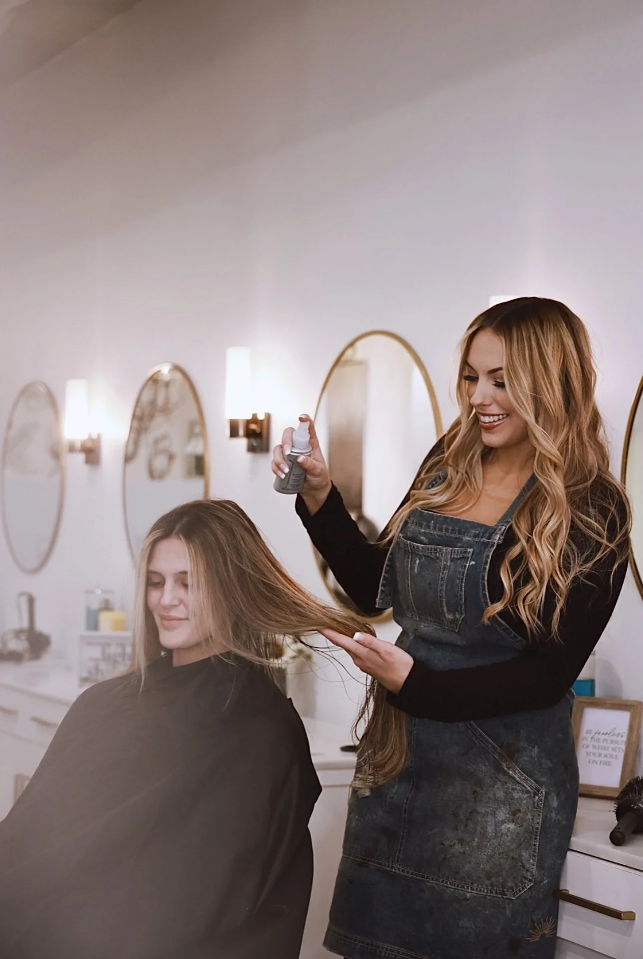 Hairdresser using a spray bottle to style a woman's long, blonde hair in a salon with oval mirrors and warm lighting.