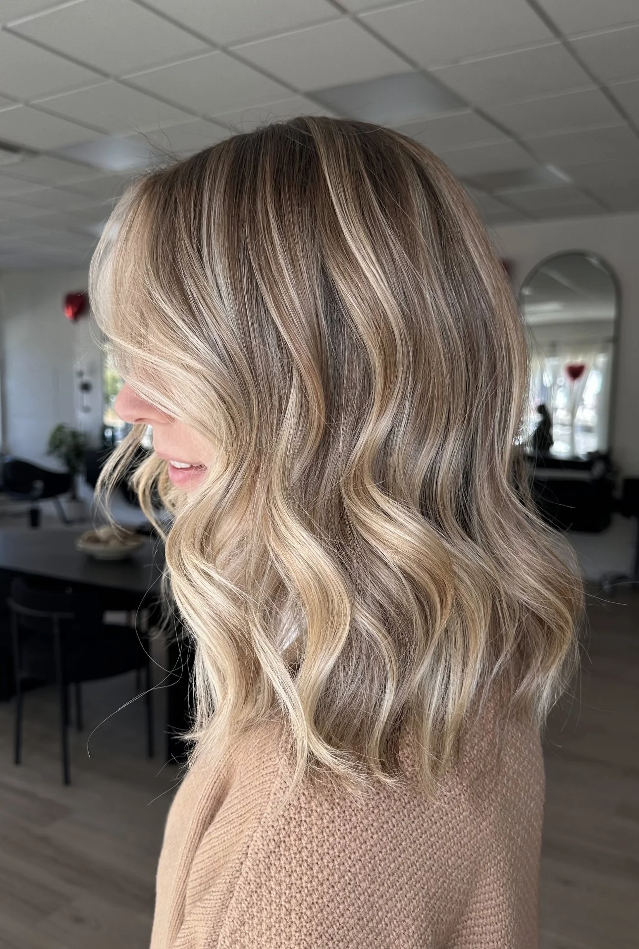 Side view of a woman with shoulder-length blonde, wavy hair in a salon, wearing a beige sweater.