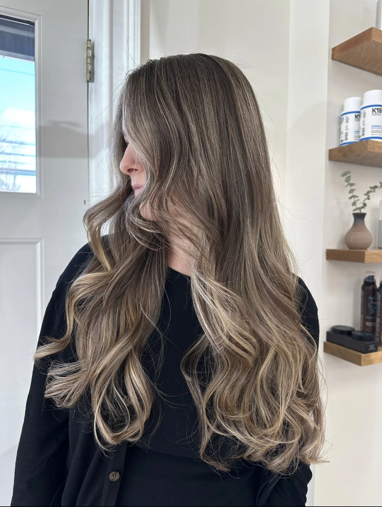 Woman with long, wavy, lived-in blonde hair  by Beauty By Kell Cart wearing a black shirt, standing indoors near a door with a window and wooden shelves with bottles and a plant.