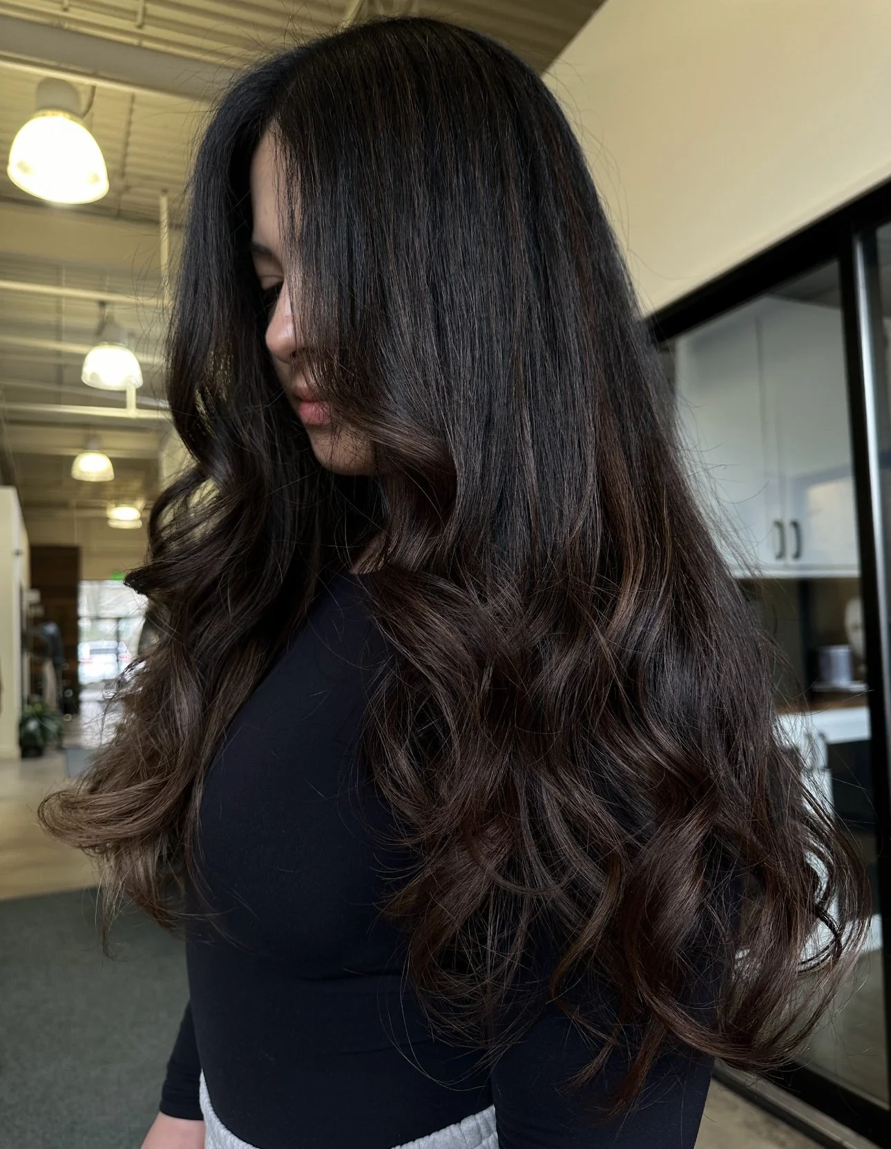 A woman with long, dark wavy hair standing indoors with modern decor and lighting.