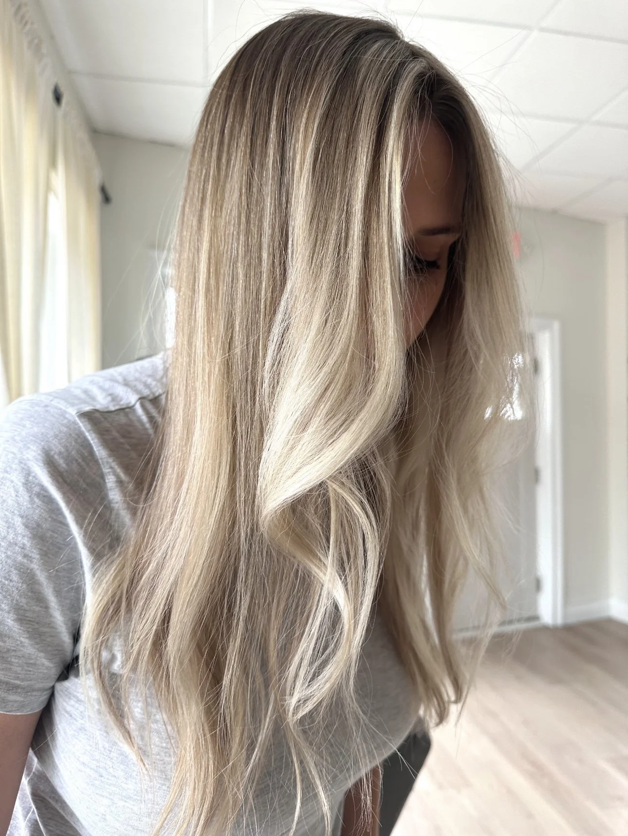 A woman with long, wavy blonde hair looking down in a bright room with white walls, window curtains, and a wooden floor.