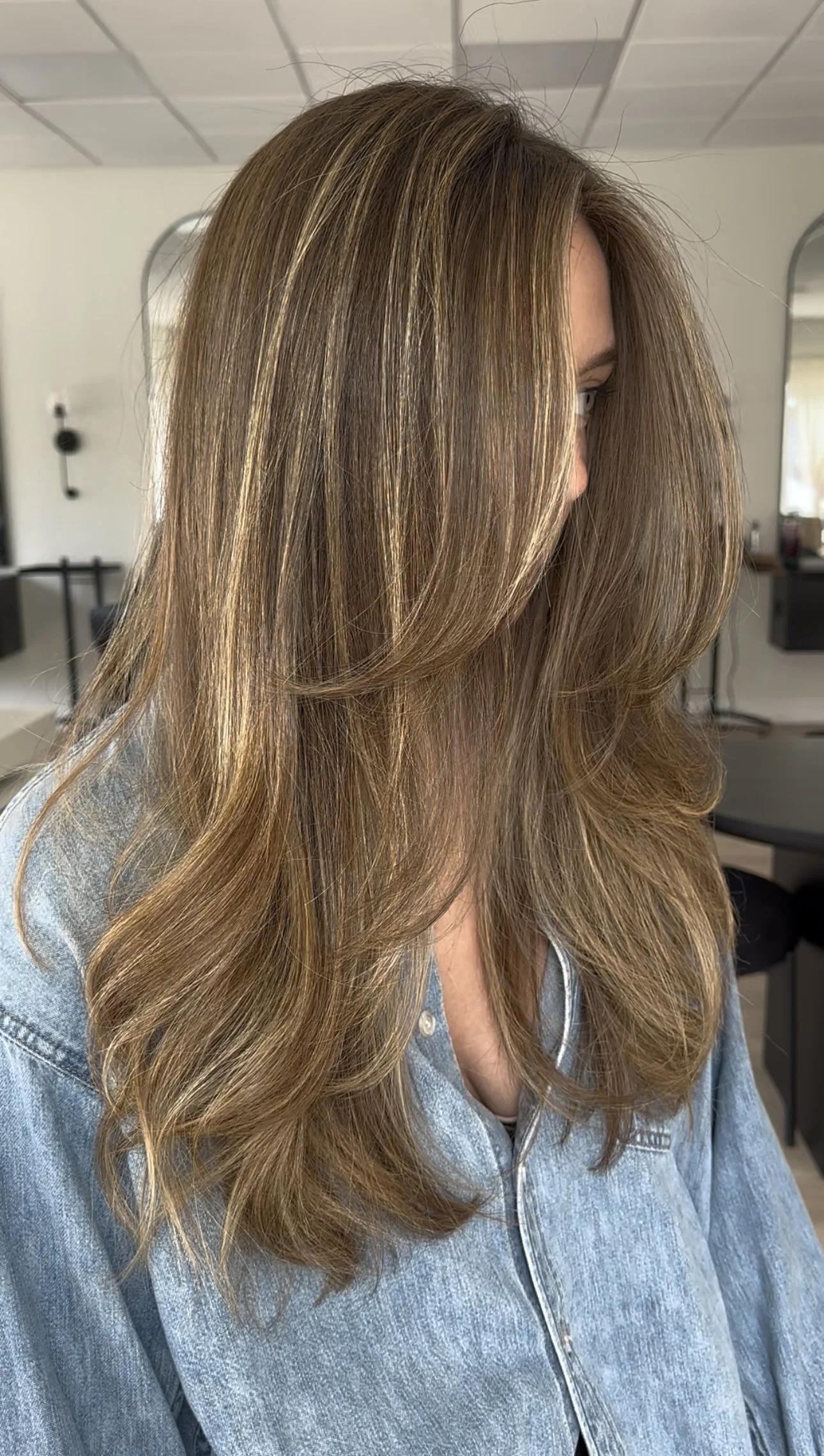A woman with long, styled light brown hair with blonde highlights, wearing a denim shirt, in a salon.