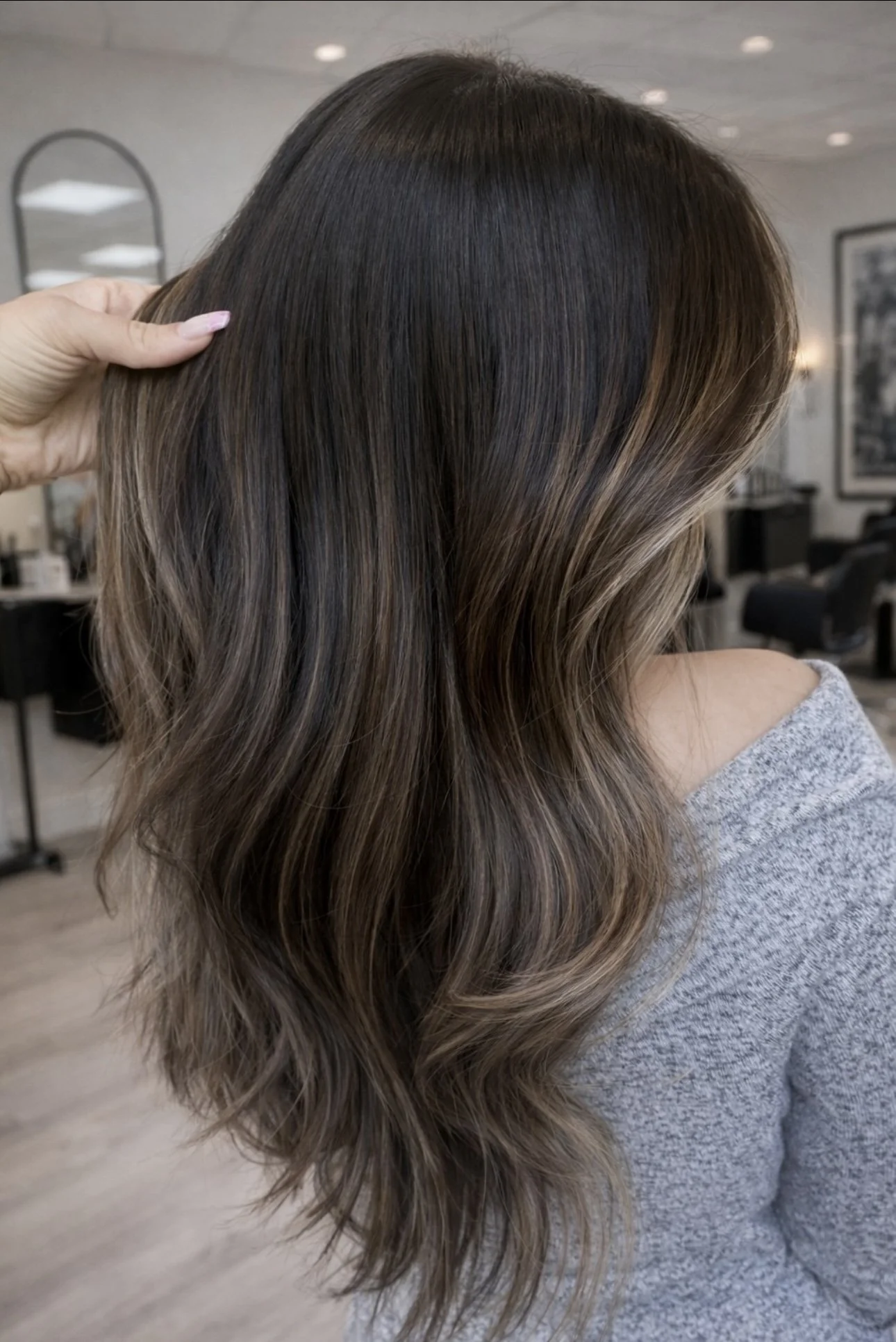 A woman with long, wavy brown hair with highlights, sitting in a hair salon, wearing a gray sweater, with a hand touching her hair.