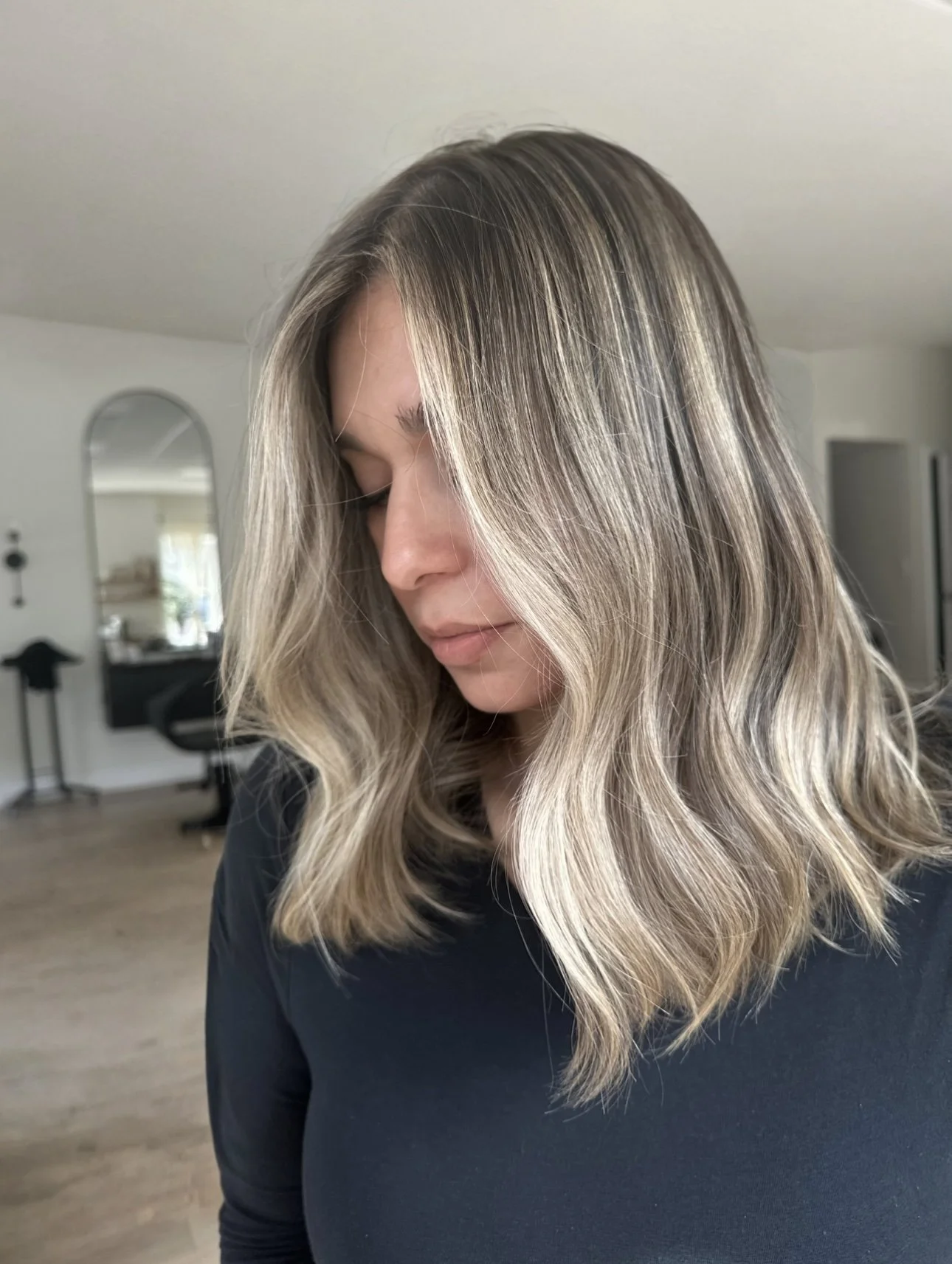 Woman with shoulder-length, wavy blonde hair looking down in a room with a mirror and some furniture in the background.