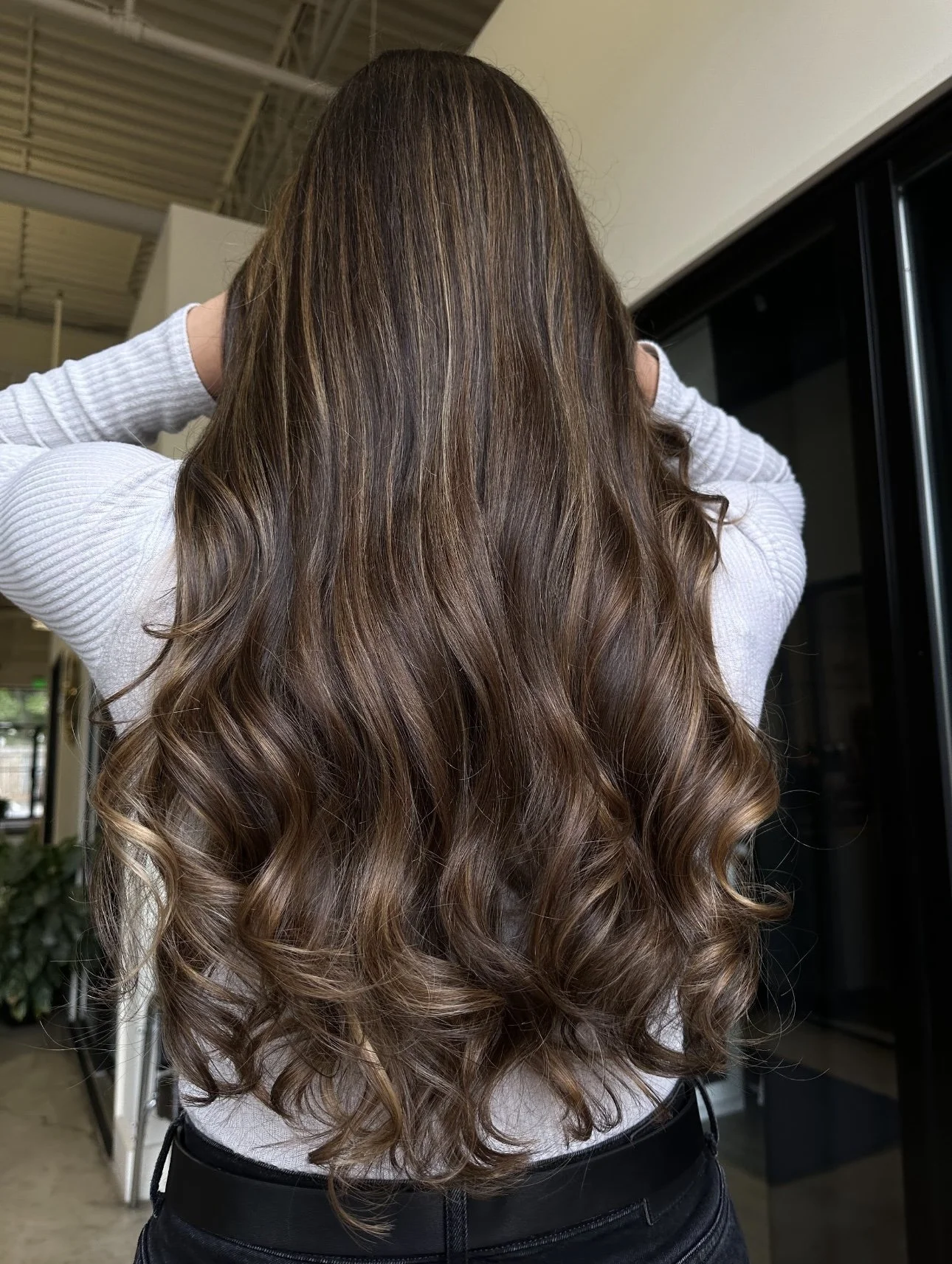 A woman with long, wavy, brown hair standing indoors, facing away from the camera, wearing a white long-sleeve top and black pants.