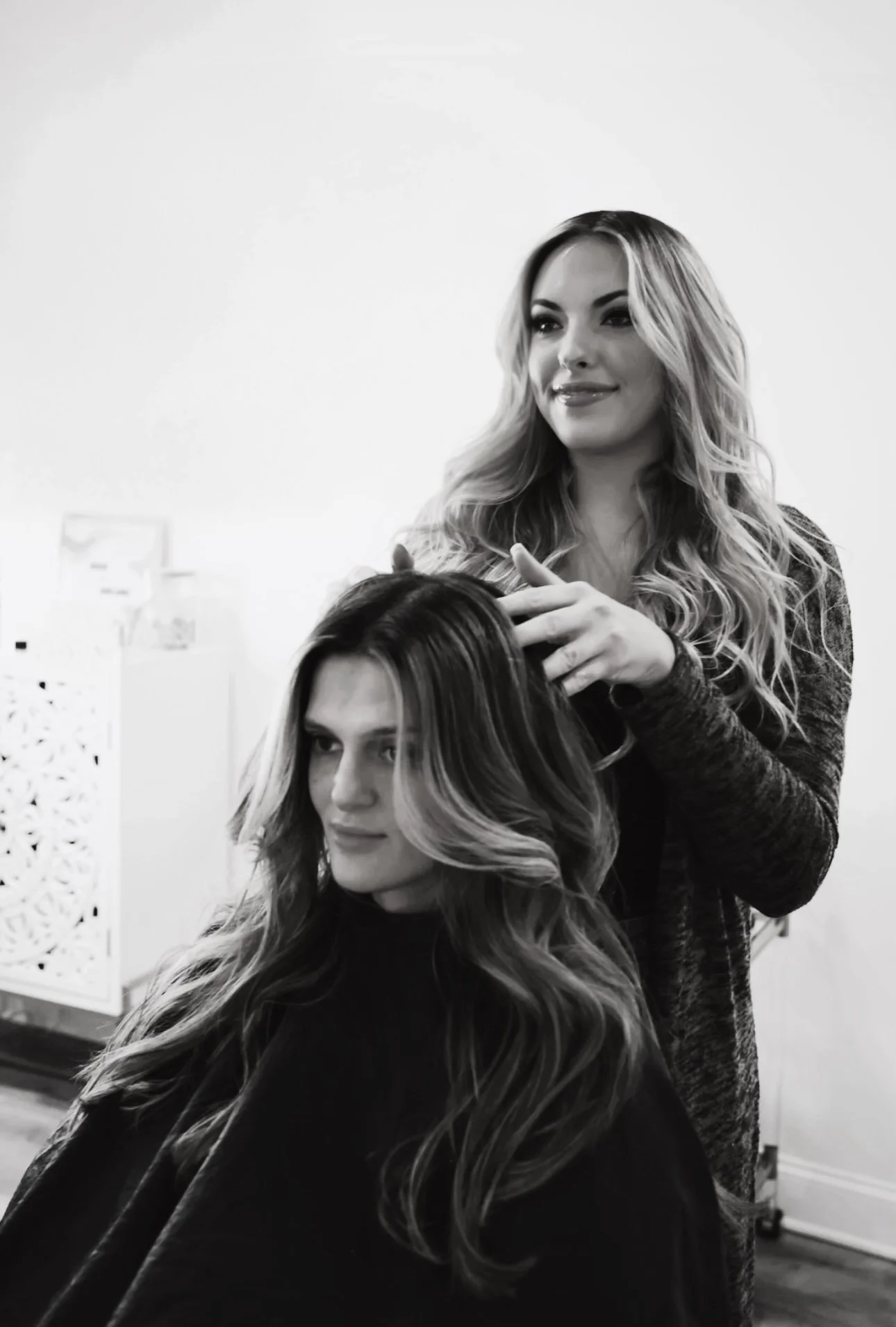 A hairstylist with long wavy blonde hair brushing the hair of a woman with long wavy hair, seated in a salon.