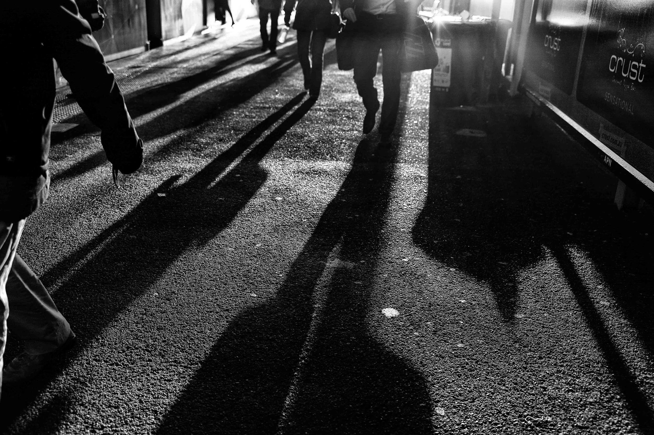 People walking on a city street during daytime, with long shadows cast on the ground from the sunlight.