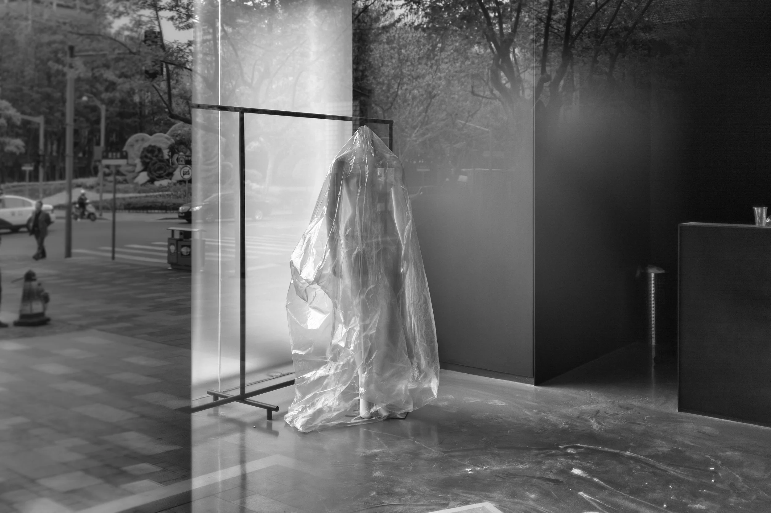 Empty indoor space with a plastic-draped sculpture or mannequin, a dark wall, a glass of water on an elevated surface, and large windows revealing a city street outside.
