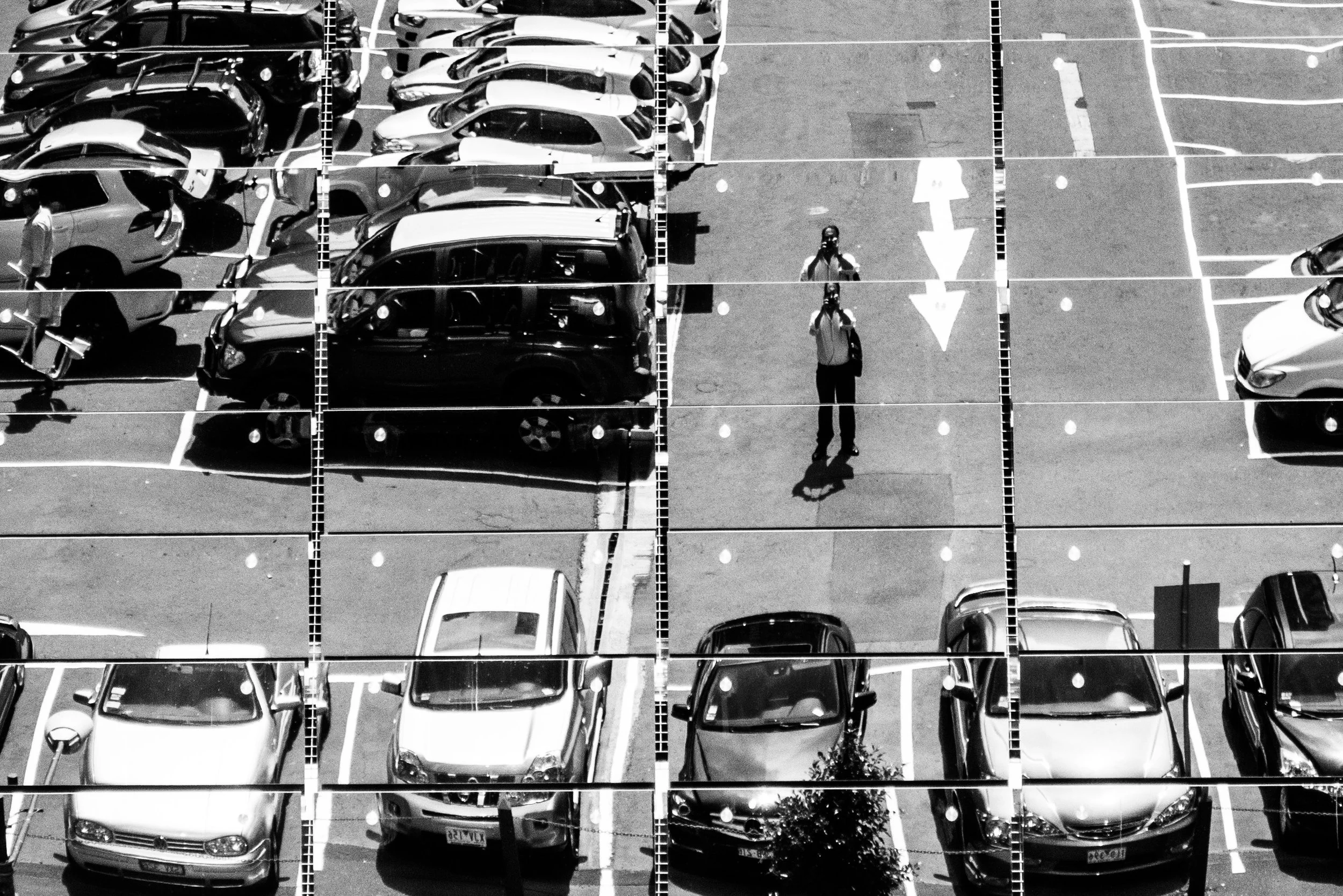 Black and white photo of a parking lot filled with cars and two people taking pictures, viewed from above.