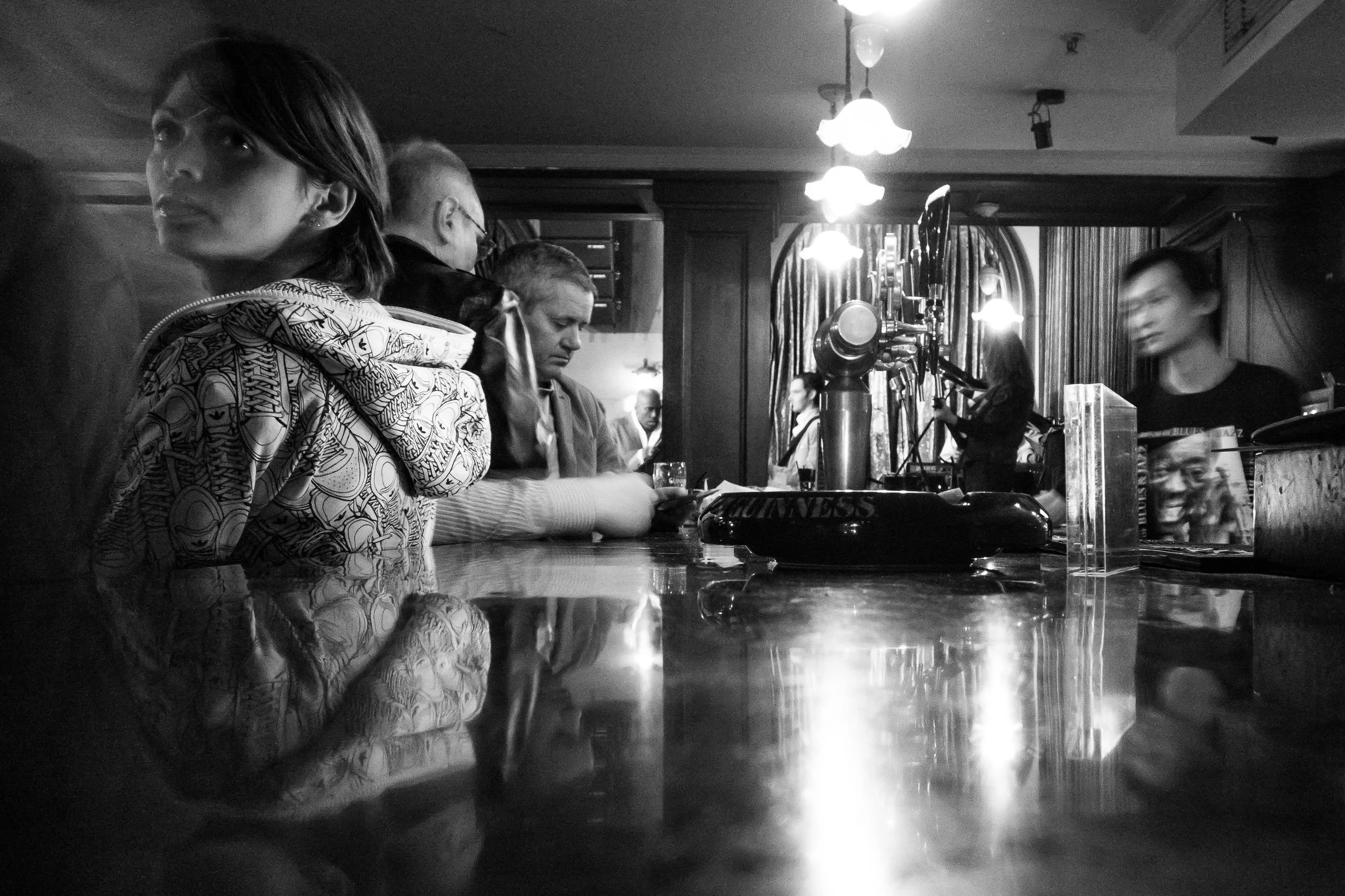 People sitting at a bar counter in a dimly lit pub, with a woman in focus on the left and musicians performing in the background.