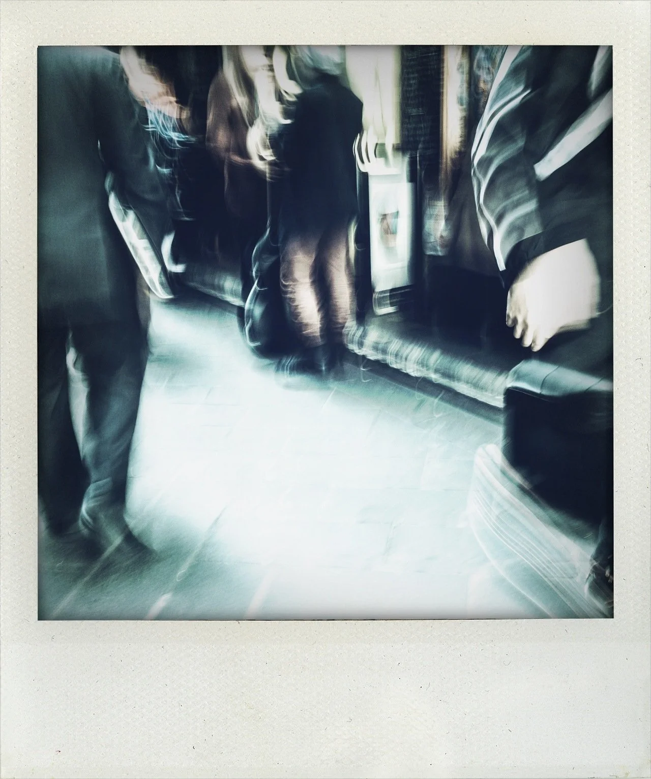 Blurred photo of people in a room, possibly a waiting area or lobby, with some seated on a bench and others standing, with a television or monitor on the wall.