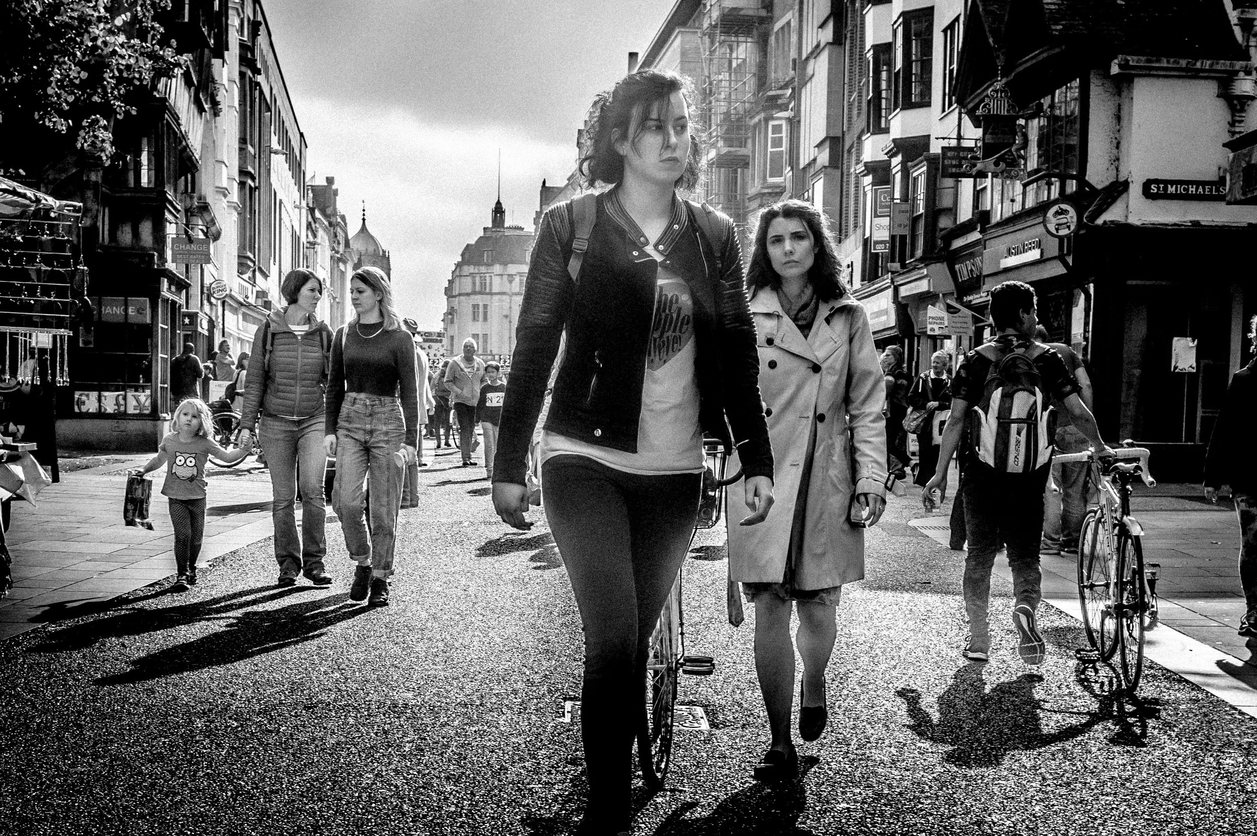 A black and white photo of a busy city street with people walking, including three women in the foreground, two of whom are walking together while the third woman walks ahead. Other pedestrians are visible in the background, along with storefronts and buildings lining the street.