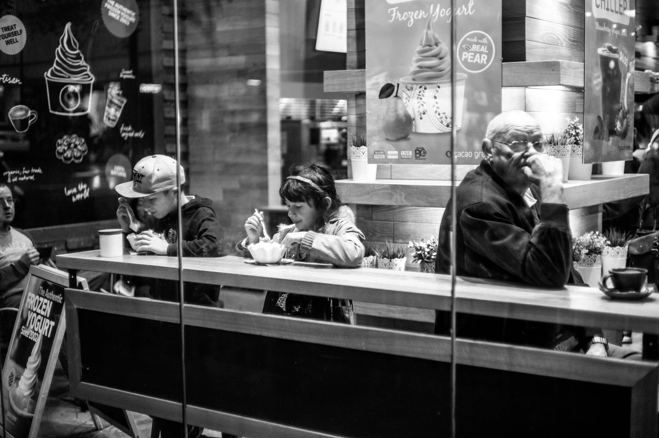 People sitting at a counter inside a cafe, seen through a window, with promotional posters and potted plants in the background.