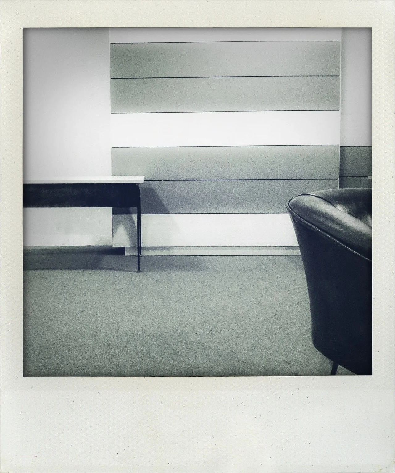 Empty office space with a black desk, a large cabinet with horizontal stripes, and a black leather chair on a gray carpeted floor.