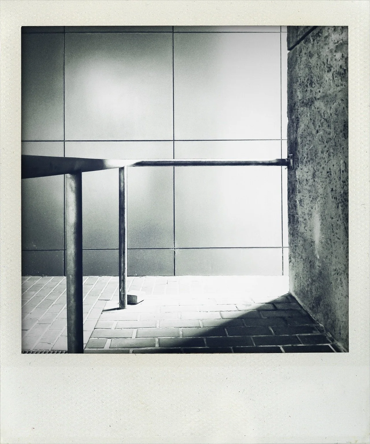 Black and white photo of a handrail and wall in an indoor space with tiled and brick flooring, illuminated by bright light.